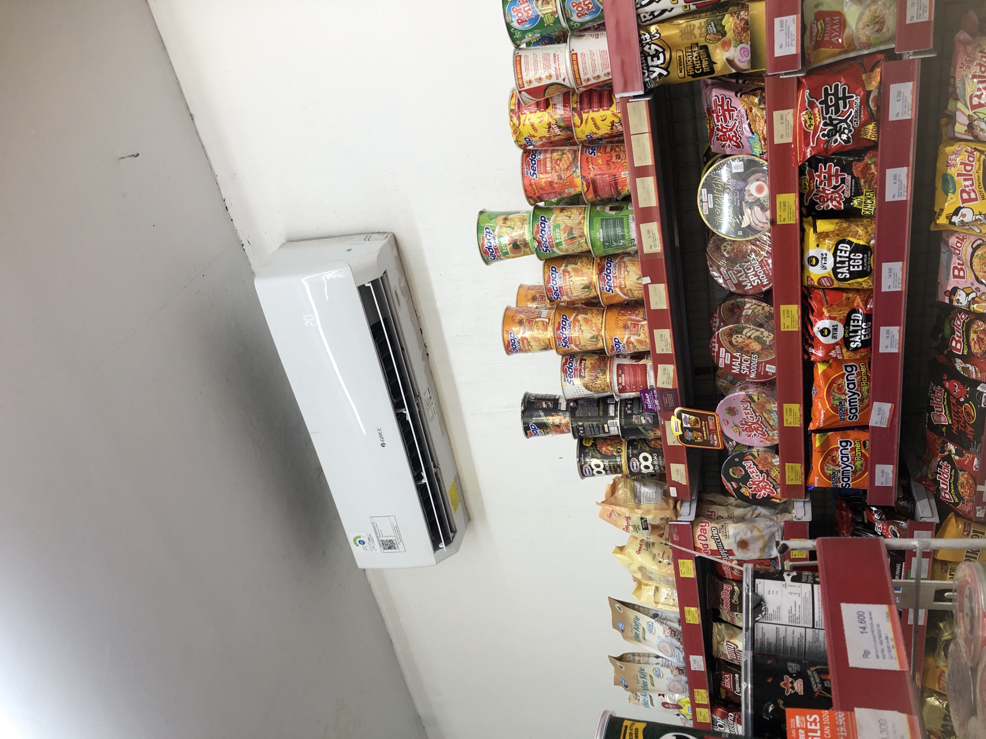 A retail store shelf is filled with various packaged food items, primarily instant noodles and coffee. The items are neatly arranged on red shelves against a white wall. An air conditioning unit is mounted on the wall above the shelves. The lighting is bright, suggesting an indoor commercial setting. The food items include brands like "Samyang," "Bulldak," "Sedap Cup," and "Good Day" coffee. Prices are displayed on small white tags in Rupiah. The scene appears to be in a store in Putat, Indonesia, a common location for such retail establishments. There are no people visible in the image.
