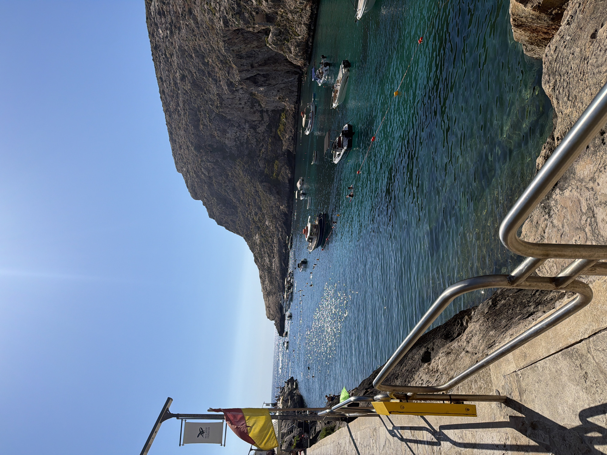 The image captures a picturesque view of Xlendi Bay in Xlendi, Malta, on a clear, sunny day. The bay is calm with crystal-clear turquoise water, dotted with numerous small boats. Several people are visible swimming and enjoying the water. The scene is framed by rocky cliffs on the right side and a stone pathway with a metal railing on the left. A flag and a banner are posted on a pole along the pathway. The sun casts shimmering reflections on the water's surface.
