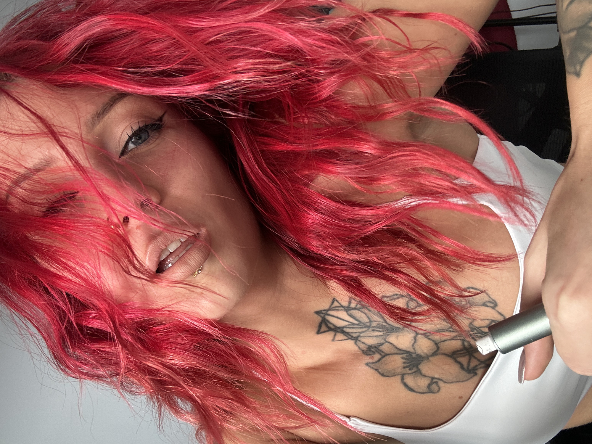 A fair-skinned woman with bright red wavy hair is positioned at an unusual angle, suggesting the photograph was taken from a top-down perspective. Her left eye is visible, framed by dark eyeliner. She has a lip piercing on the lower left side of her mouth. She is wearing a white top, exposing part of her chest, where a large tattoo of a floral design with geometric elements is visible. A hand holding a cylindrical metallic object is positioned close to her chest. The background suggests an interior setting with elements of black furniture visible. There is no information in the image indicating it was taken in North Kuta, Indonesia.