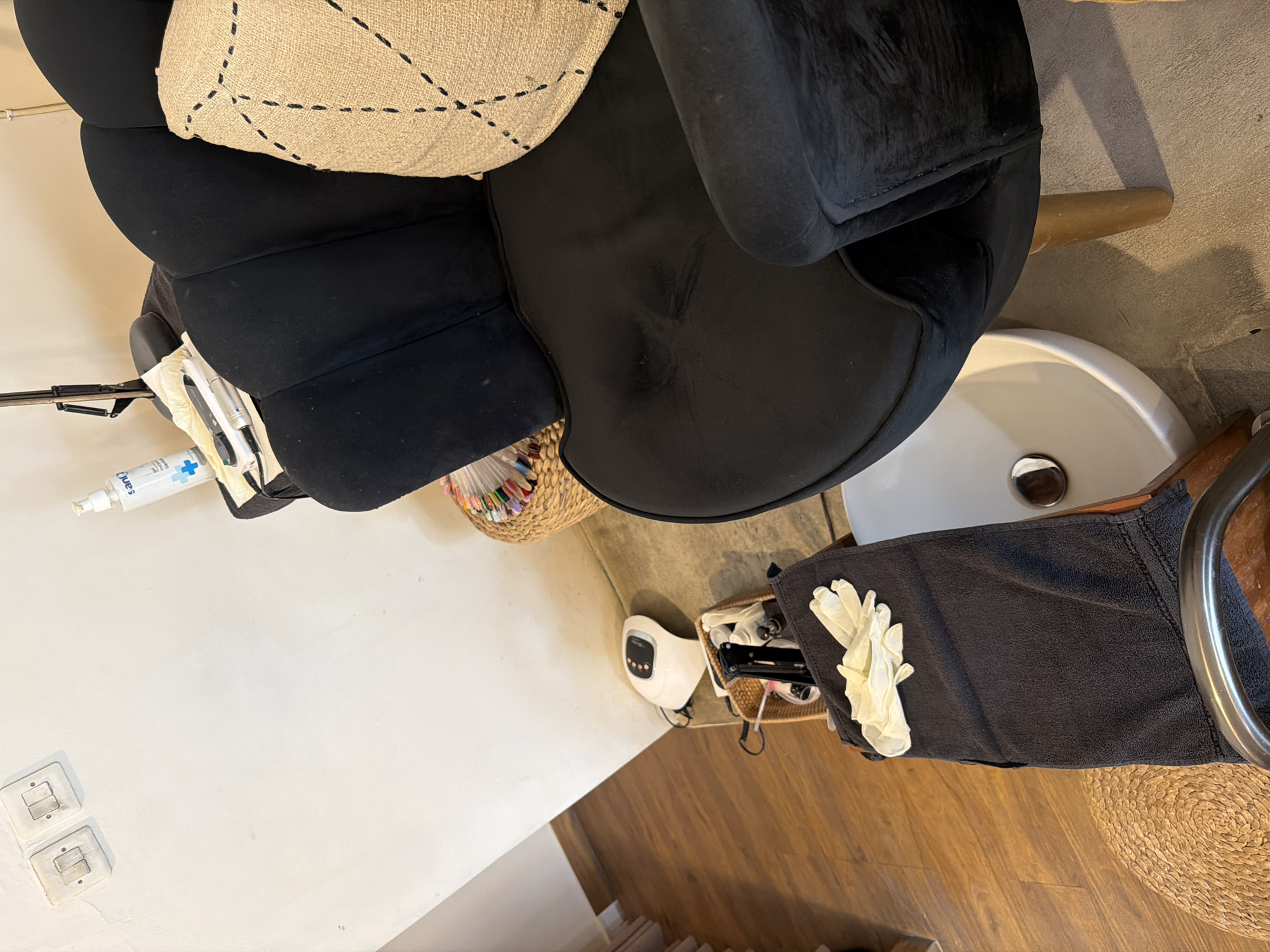 A black velvet armchair with a beige patterned throw pillow sits next to a white foot spa bowl. A dark grey towel with a pair of white latex gloves is draped over the armrest of a chair in the foreground. To the left of the armchair, on a wooden floor, is a white foot bath machine. Various manicure supplies are visible, including a wicker basket filled with nail polish bottles and a fan of colored nail tips displayed in a woven container. In the background, a white wall features two light switches. A bottle of hand sanitizer sits on a shelf near a lamp. The setting appears to be a nail salon in North Kuta, Indonesia.