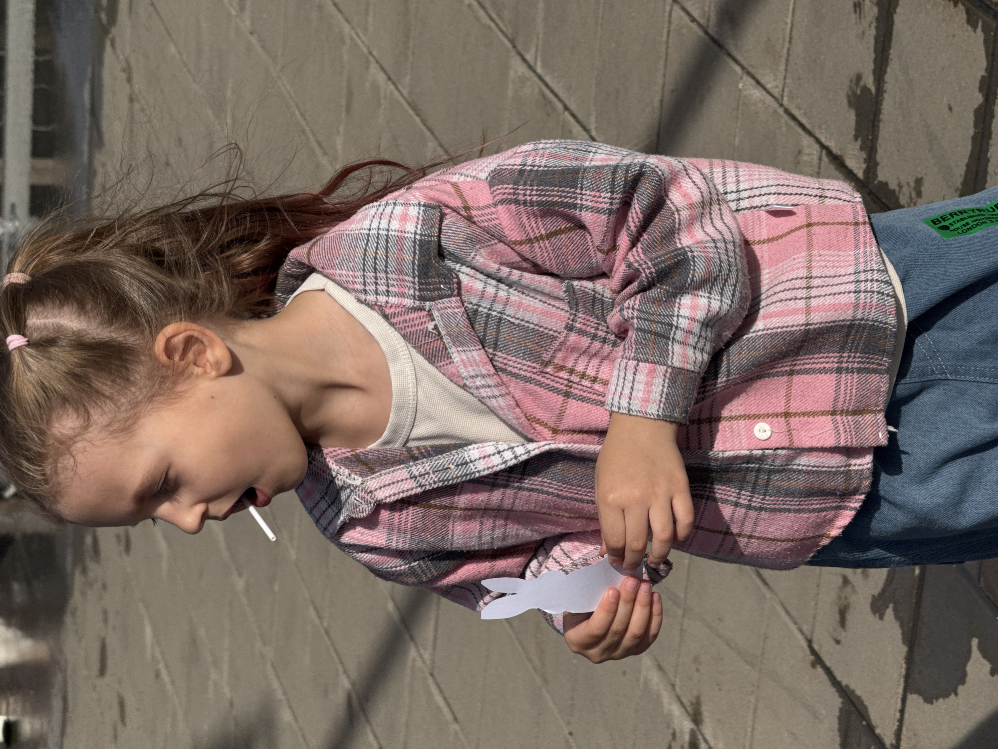 A young girl stands on a gray brick surface, holding a white paper cutout of a rabbit. She is wearing a pink and gray plaid shirt over a white top, and denim jeans. A white lollipop stick protrudes from her mouth. A green tag on the jeans reads "BERRYRUB," "ESTABLISHED," "HOUSE HO," "LONDON," and "S." The photo is taken during the daytime. The location is not discernible from this image alone.