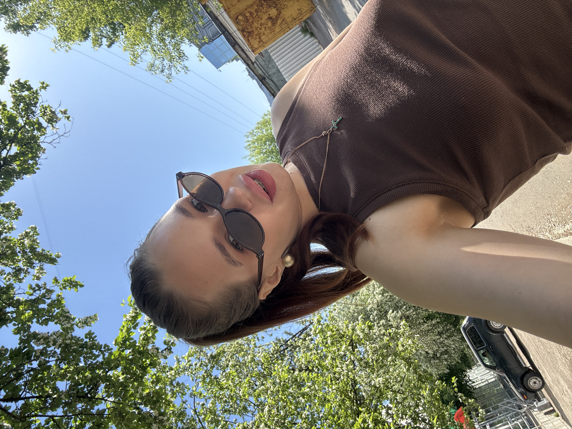 The image depicts a woman with brown hair and sunglasses wearing a brown tank top. She is outdoors, with trees and a blue sky visible in the background. There is a car parked nearby and buildings in the distance, which indicates the location is in a city. Based on the prompt, this is likely to be in Chaikovsky, Russia. The picture is taken from a low angle, looking up at the woman.