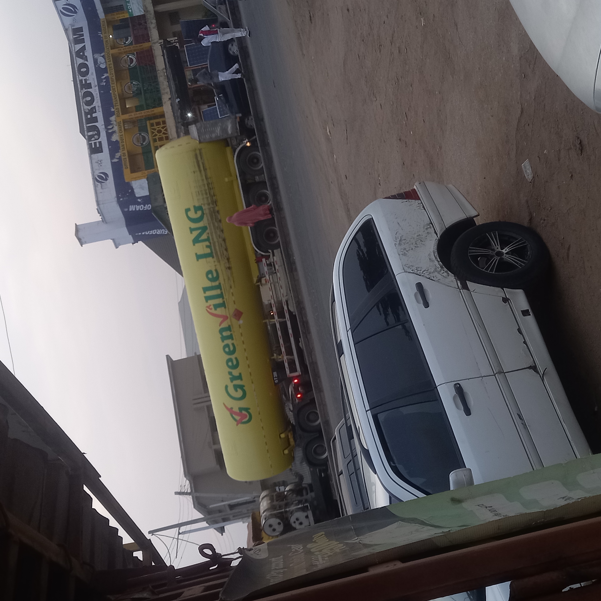The image shows a street scene in what appears to be Dadi Riba, Nigeria. A large, yellow "Greenville LNG" tanker truck is parked on the side of the road. Beside the truck is a white passenger car. Several people, including one wearing a red dress, are standing near the truck. In the background, there is a building with a sign that says "EUROFOAM" and the "OFFICE OF YAN DOYA CHAPEL ROAD TRANSPORT EMPLOYER'S ASSOCIATION OF NIGERIA." The ground is dirt and gravel. Overhead is a partial view of a makeshift awning or structure.