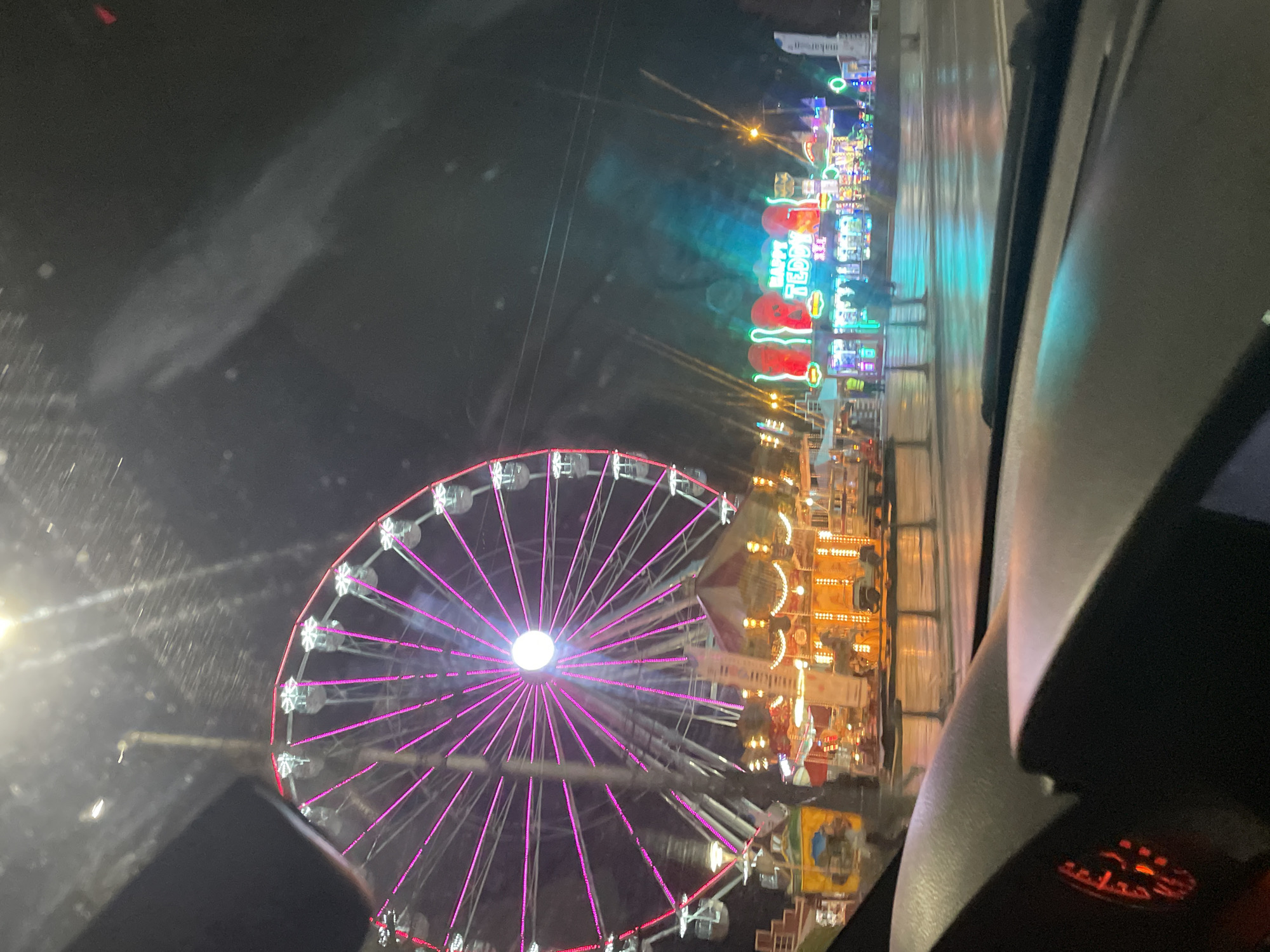The image shows a night scene taken from inside a vehicle, possibly a car. The primary focus is a large Ferris wheel illuminated with pink lights. In the background, there's a fair or carnival scene with various lit-up structures, including one with the words "HAPPY TEDDY". The overall scene is bright due to the artificial lights of the Ferris wheel and carnival attractions. The windshield of the vehicle is visible, and there appear to be raindrops or moisture on the glass. The interior of the vehicle, including parts of the dashboard and steering wheel, are also visible. The location cannot be determined as Sofia, Bulgaria.