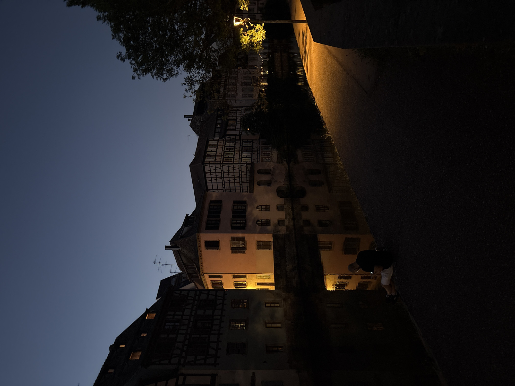 The image shows a nighttime scene in Strasbourg, France, likely in the Petite France district. Half-timbered buildings line a dark waterway. A sidewalk runs alongside the water, illuminated by a street lamp. A single person stands on the sidewalk, bending over slightly and looking down, possibly at a device. The sky is a dark blue, transitioning to black at the top of the frame. The buildings are reflected in the water, creating a mirror image.