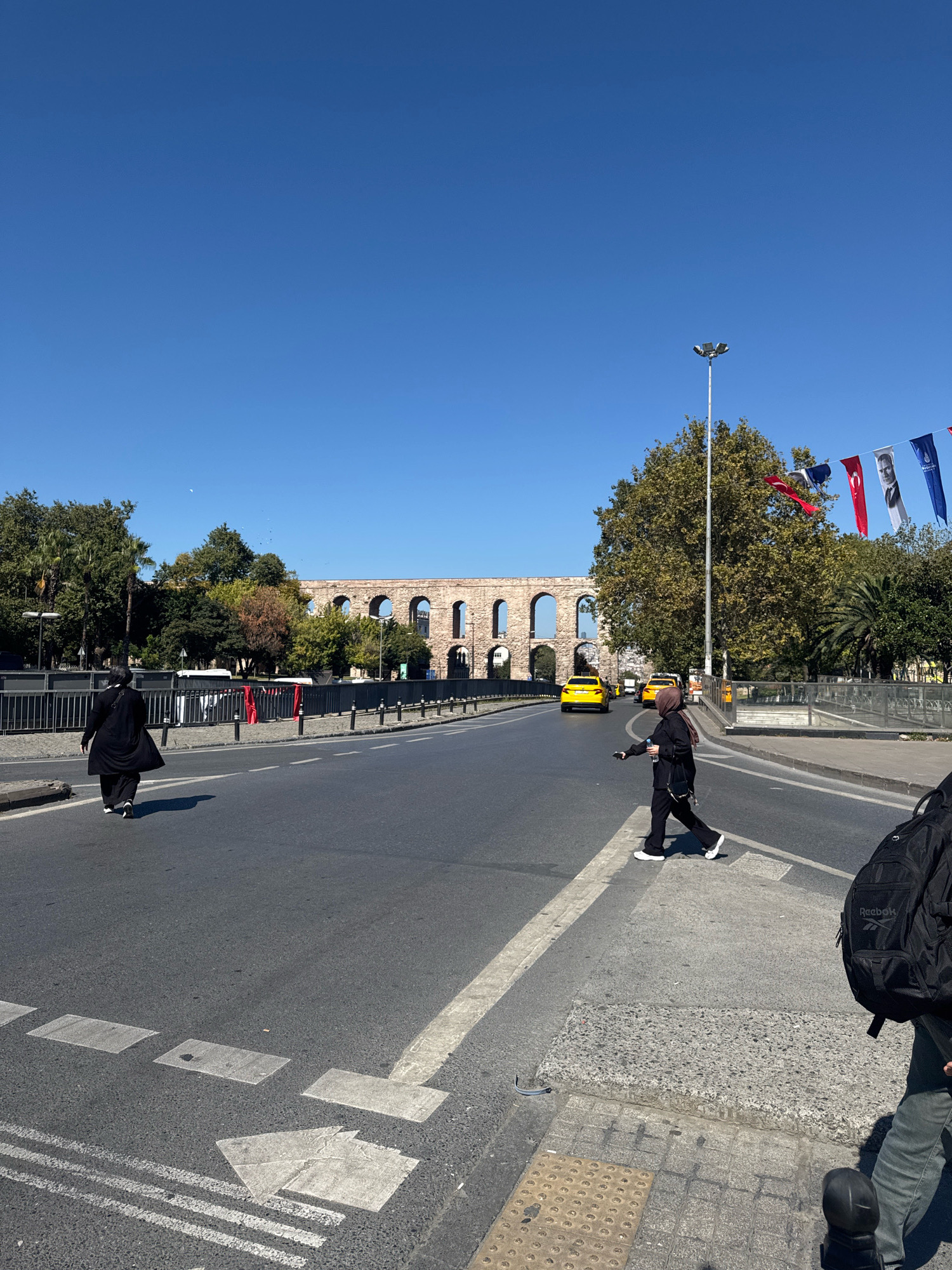 In Istanbul, Turkey, on a clear day with a bright blue sky, a street scene unfolds. An ancient stone aqueduct, characterized by its arched structure, stretches across the background. In the mid-ground, a tree-lined street features several cars, including two yellow taxis, and a lamppost. Decorative flags with portraits hang from a wire stretched between poles. Two women are visible; one, wearing a black abaya and hijab, walks on the left side of the road, while another, also in black with a hijab, crosses the street at a designated crosswalk. The foreground shows a portion of the road with crosswalk markings and a sidewalk with tactile paving. The overall environment suggests a bustling urban area with historical significance.