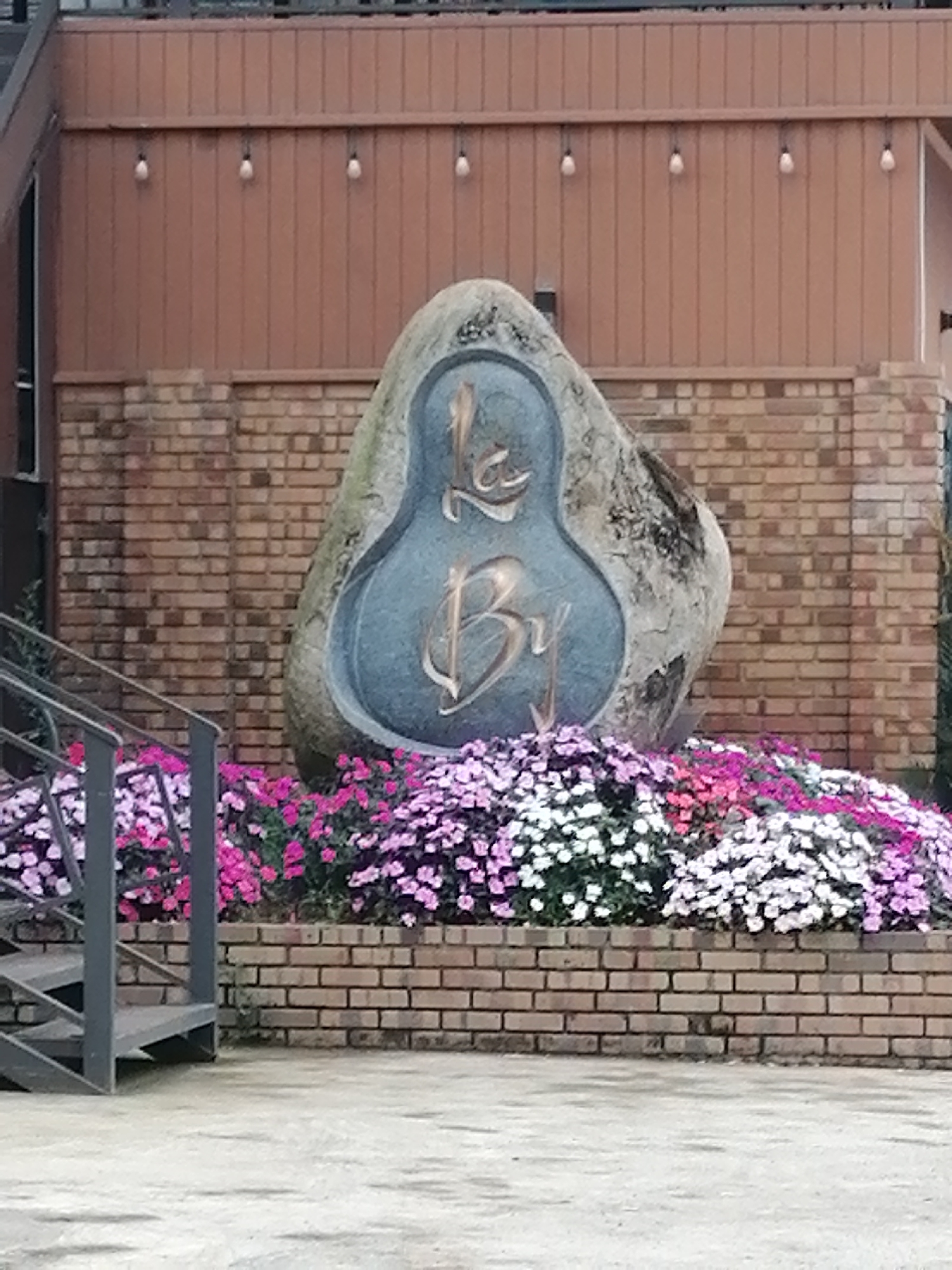 The image displays an outdoor scene, likely a public space or entrance in Lạc Dương, Vietnam. At the center is a large, irregularly shaped natural stone monument, roughly pear-shaped, with a prominent inscription carved into its surface. The stylized script on the stone clearly spells out "Lạc Dương," with "Lạc" appearing above "Dương" within an oval, recessed area that is a muted blue-gray color. The letters themselves are in a golden or bronze tone.
In front of the stone monument, a dense and colorful flower bed extends across the width of the frame, filled with numerous small blossoms in vibrant shades of pink, purple, and white. This flower bed is bordered by a low, light brown brick wall. To the left, a dark gray metal staircase with open risers and diagonal supports descends towards the foreground.
Behind the stone and the flower bed, another brick wall rises, topped by a light brown wooden panel wall. Several individual string lights or bare bulbs are evenly spaced and hang from this wooden wall. The ground in the immediate foreground is a plain, light gray concrete surface. The lighting suggests an overcast or cloudy daytime, as there are no strong shadows and the illumination is soft and diffuse. The scene conveys a sense of tranquility and a well-maintained environment.