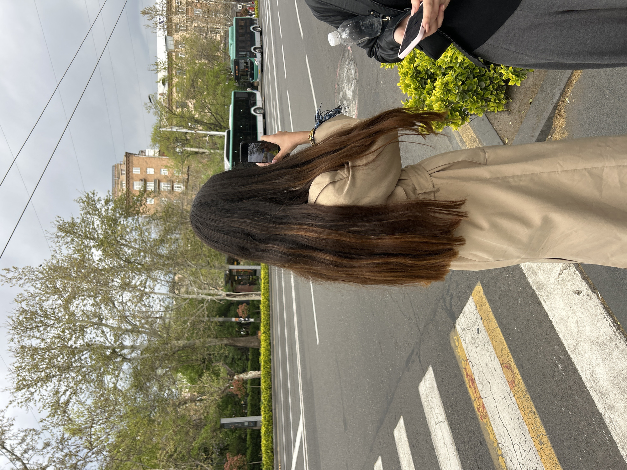 A woman with long, dark hair takes a photo with her phone on a crosswalk in Yerevan, Armenia. She is wearing a beige coat. Another person stands next to her, holding a phone and a water bottle. Behind them, buses line the street. Trees and buildings are visible in the background, under a cloudy sky.