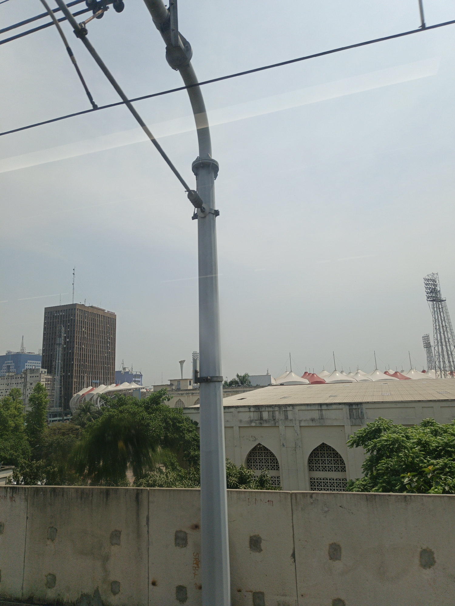 The image, taken from what appears to be a moving vehicle, shows a cityscape in Dhaka, Bangladesh. An overhead power line structure is prominent in the foreground. Behind it, a tall, brown office building stands out. To the right of the office building, part of the Bangabandhu National Stadium is visible, characterized by its white and red peaked roof tents and floodlights. A concrete barrier separates the viewer's perspective from the immediate scenery. The sky is overcast.