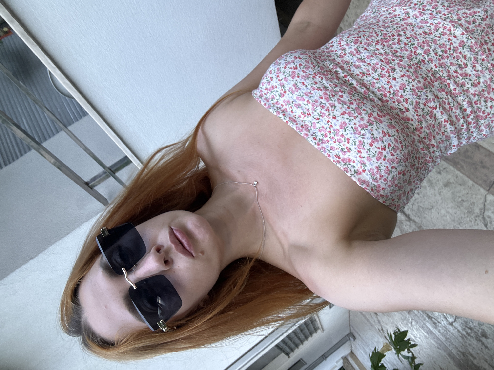 A young woman with auburn hair and sunglasses is posing for a photo outdoors. She is wearing a strapless floral-print dress and a delicate silver necklace. The background shows a white wall and some outdoor flooring. The image appears to have been taken in Budva, Montenegro, specifically near a residential building with a metallic fence and some greenery.