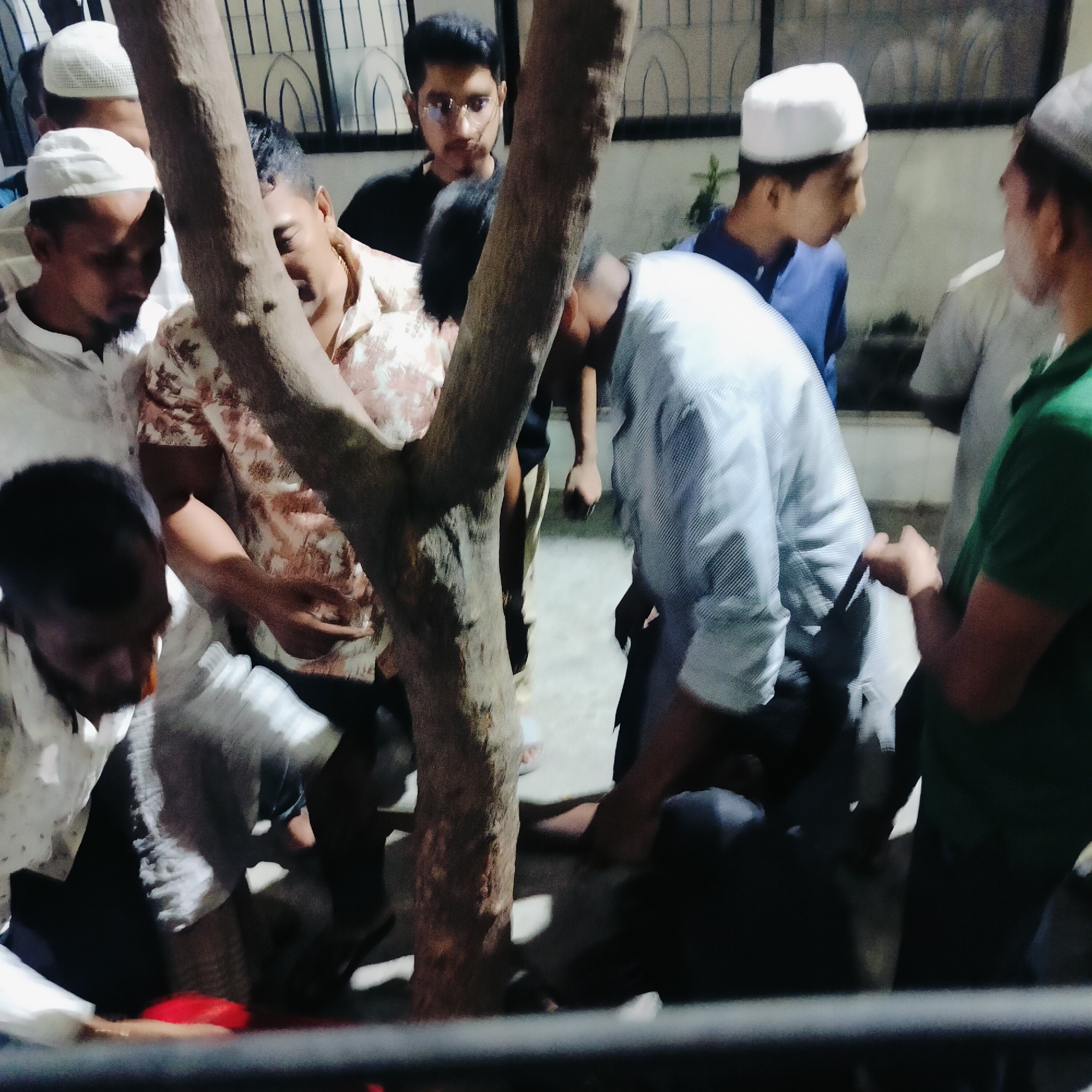 A group of men, several wearing white skullcaps, are gathered around what appears to be a fallen object or person near a tree. The location is potentially Dhaka, Bangladesh, based on the apparent ethnicity of the people. The image is taken at night or in a dimly lit environment, with some figures facing down towards the object of interest. Some are wearing casual clothing, while others are in more traditional attire. There is a building in the background with barred windows.