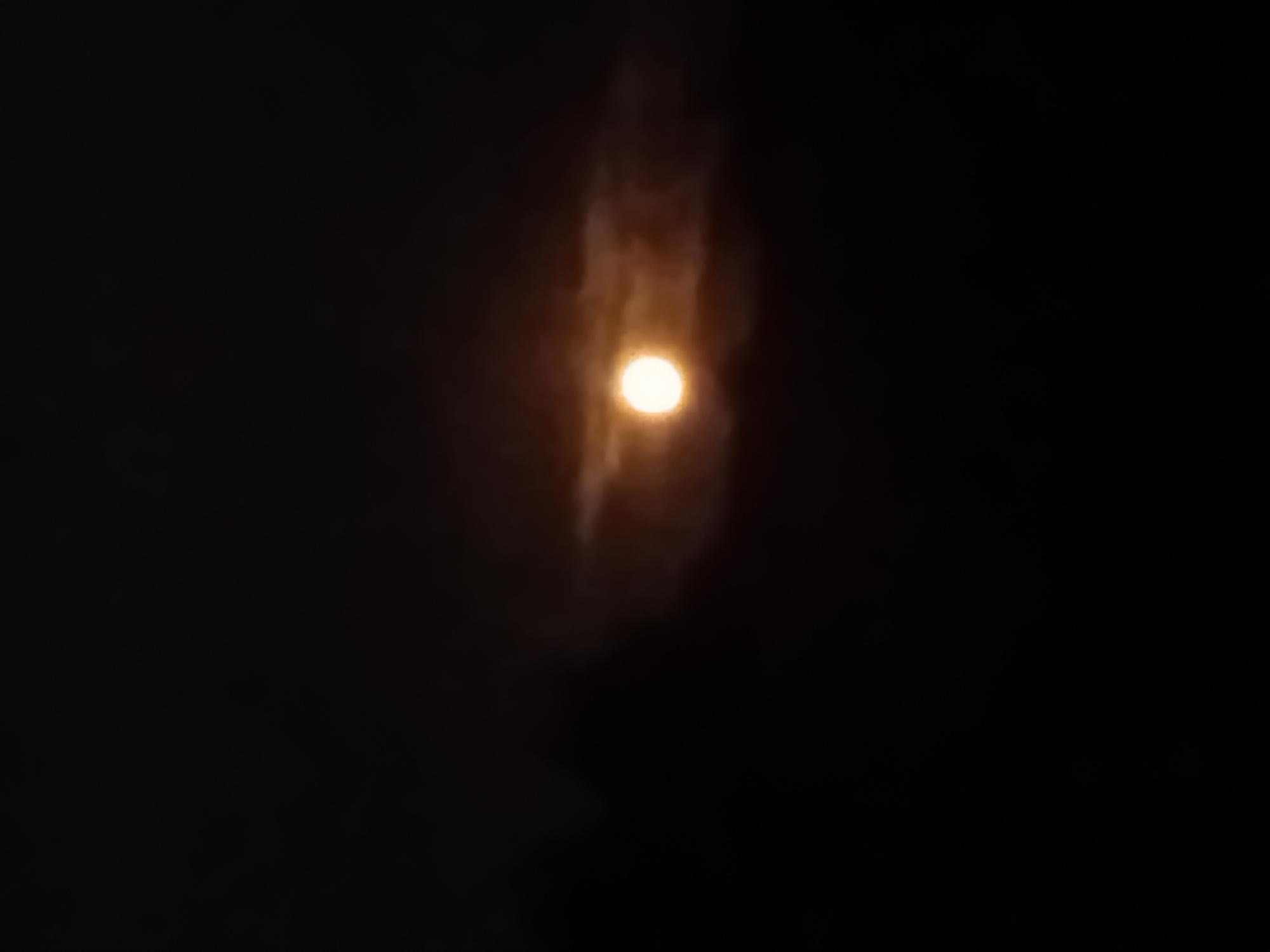 A full moon is partially obscured by clouds against a dark sky. The moon is bright and appears as a yellow-orange orb, with some lighter clouds glowing around it. The rest of the image is predominantly black, indicating nighttime. The location is the city of Verkhniachka, Ukraine. There are no people or objects visible in the image, only the celestial body and atmospheric conditions.