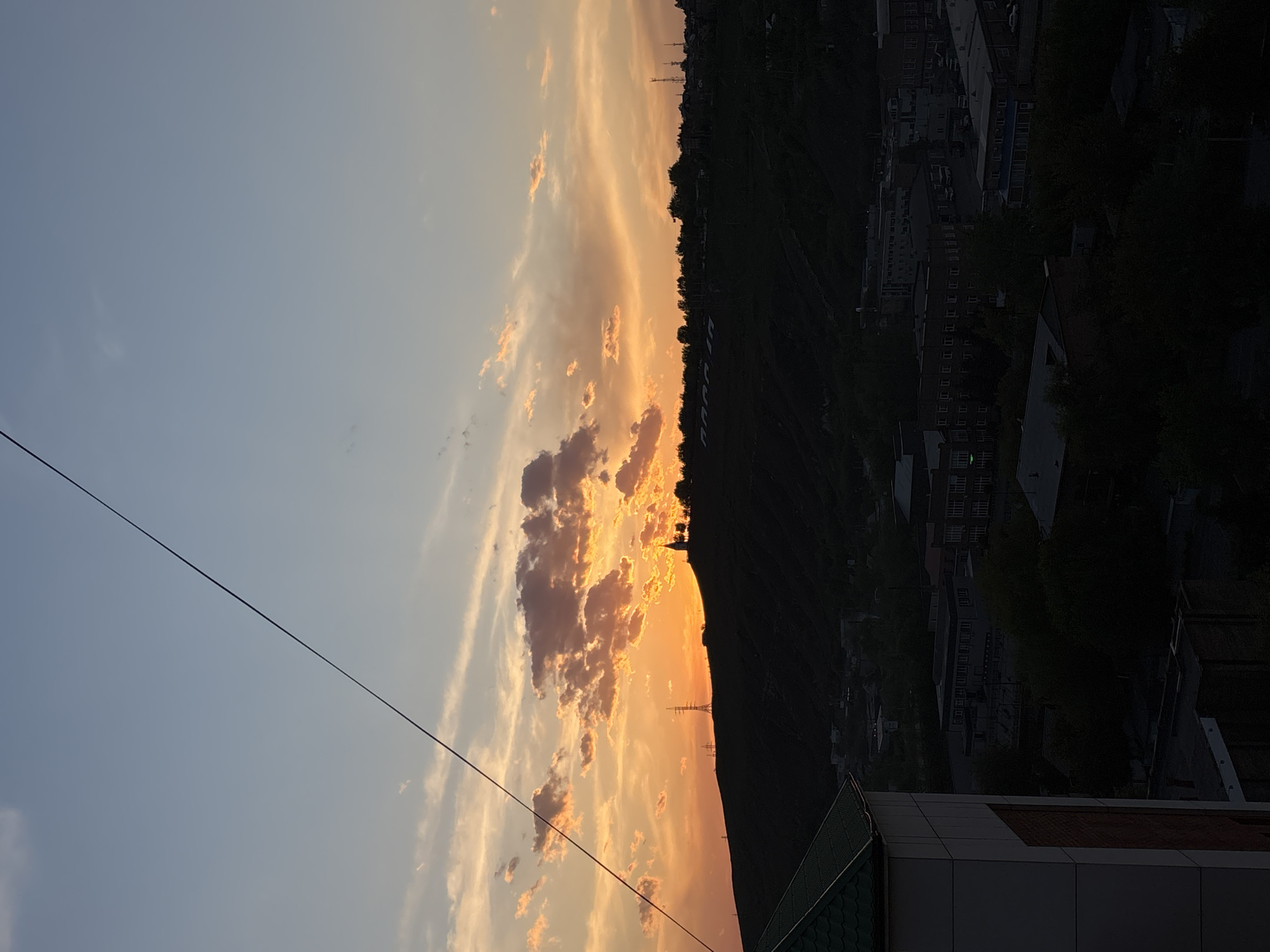 The image depicts a sunset in what appears to be a cityscape. The sky transitions from a light blue on the left to a vibrant orange and yellow on the right, filled with clouds. Silhouetted against the sunset is a dark, elevated land mass that appears to be a hill or mountain. A line of trees and some buildings are visible on the crest of the hill.
The lower right corner of the image shows a building with a green roof.
The presence of the hill suggests the city is likely Krasnoyarsk, Russia, as it is known for being situated near hills and mountains. The angle of the photograph is taken downward.