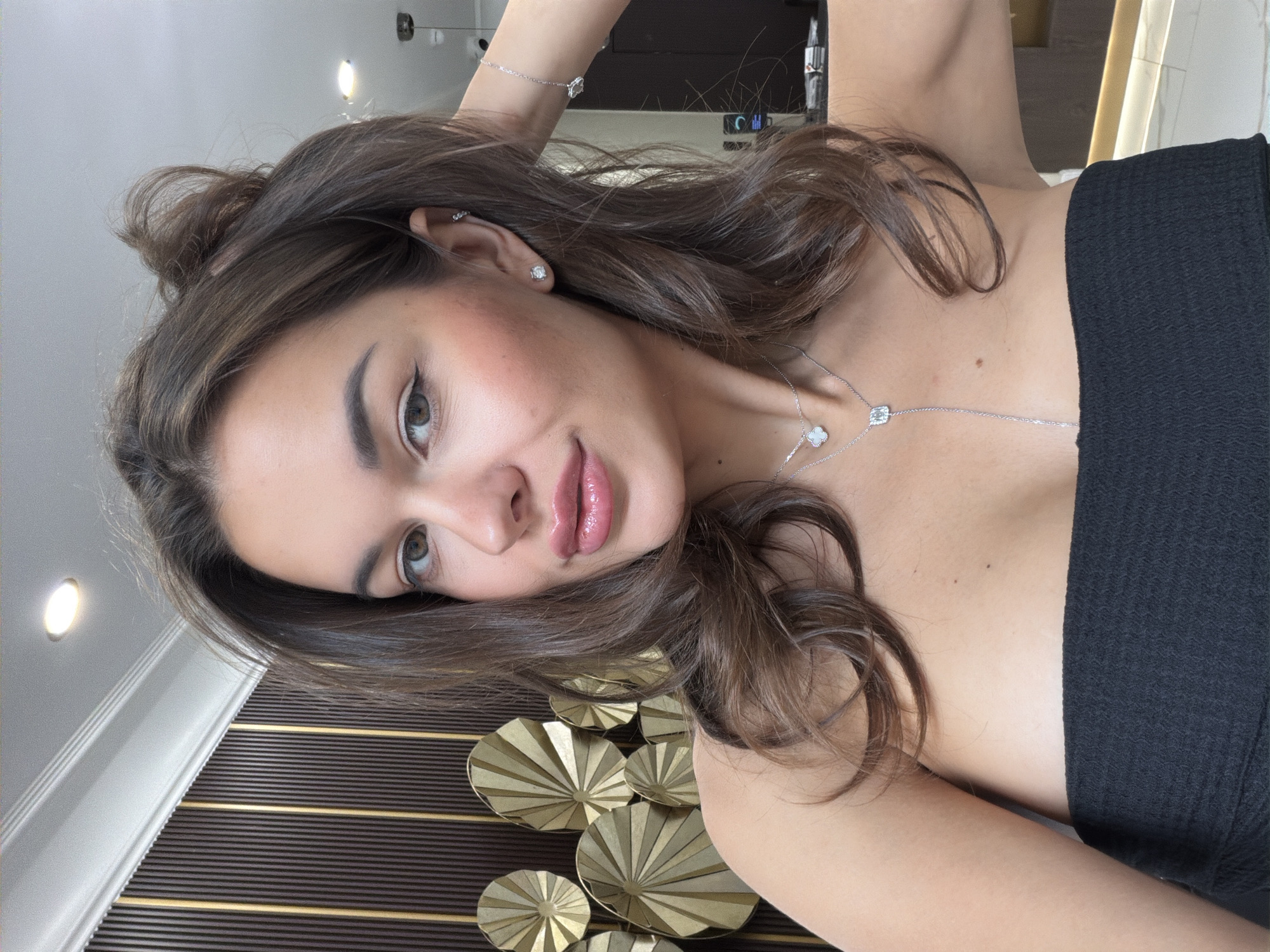 A woman with long brown hair is lying down, facing the camera. She is wearing a black top and jewelry, including earrings, a bracelet, and a necklace. The background includes a decorative wall with golden fan-like elements. The image does not contain enough information to place it in the city of Krasnoyarsk, Russia.