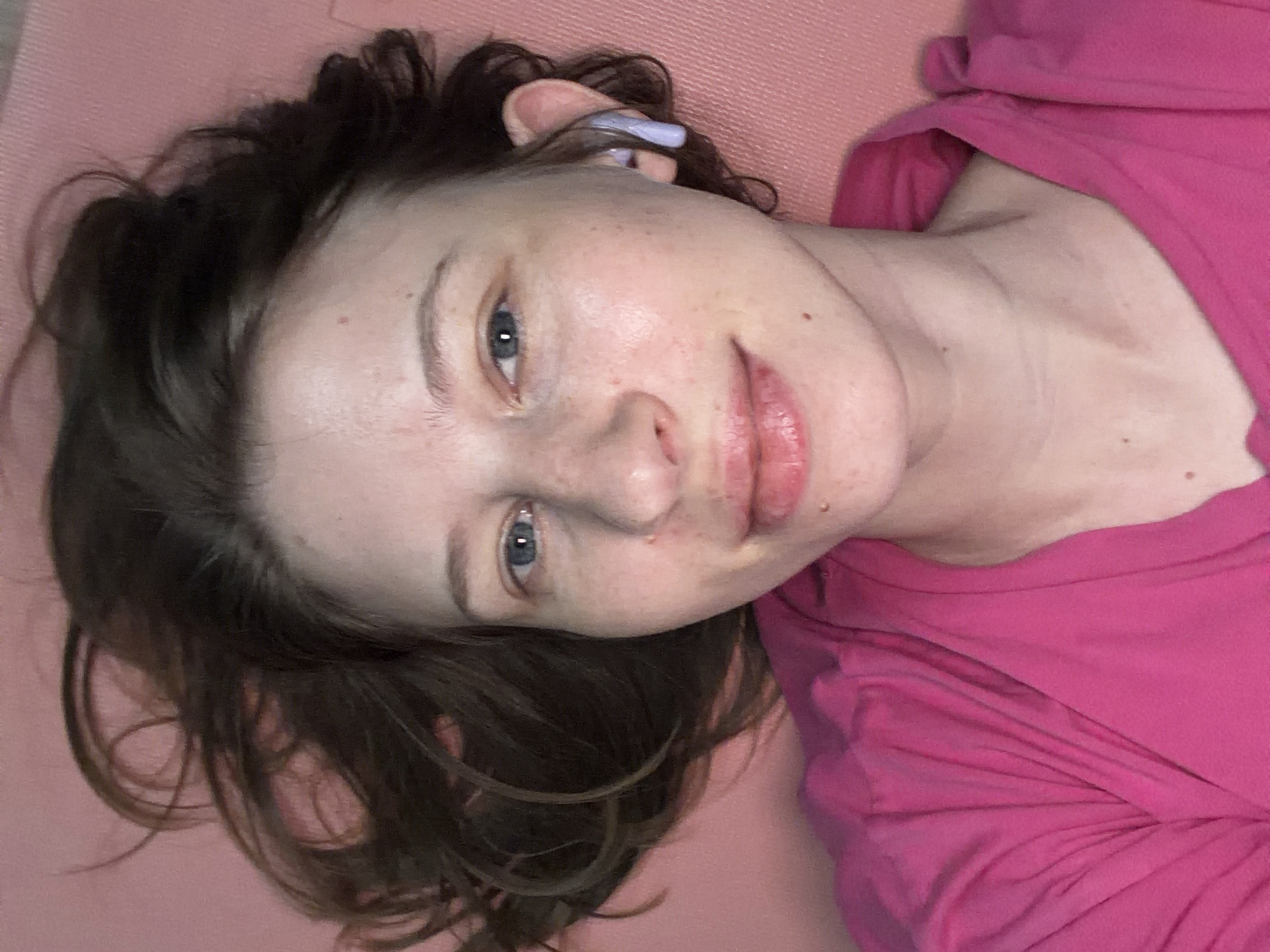 A person with brown hair and blue eyes, wearing a pink shirt and earbuds, lies on a pink mat. The image is a close-up, overhead shot of their face and upper body. There are no discernible landmarks or environmental details to indicate a specific location within Dongducheon-si, South Korea.