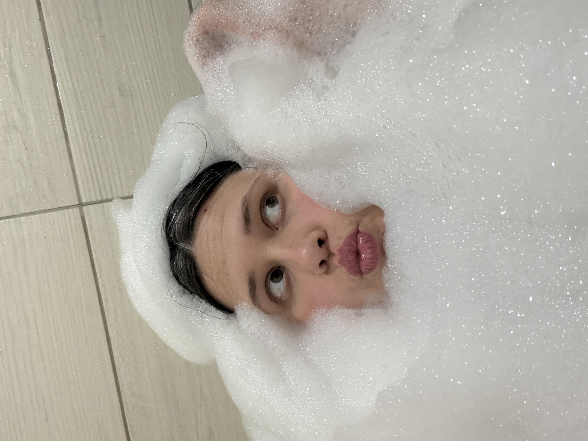 A young woman is lying in a bathtub filled with soap bubbles. The bubbles surround her face, leaving her nose, mouth, and eyes visible. She is looking upwards and slightly to the left. The background is a tiled wall and floor. The location is likely a bathroom, but there are no defining features to identify it as Chaikovsky, Russia.
