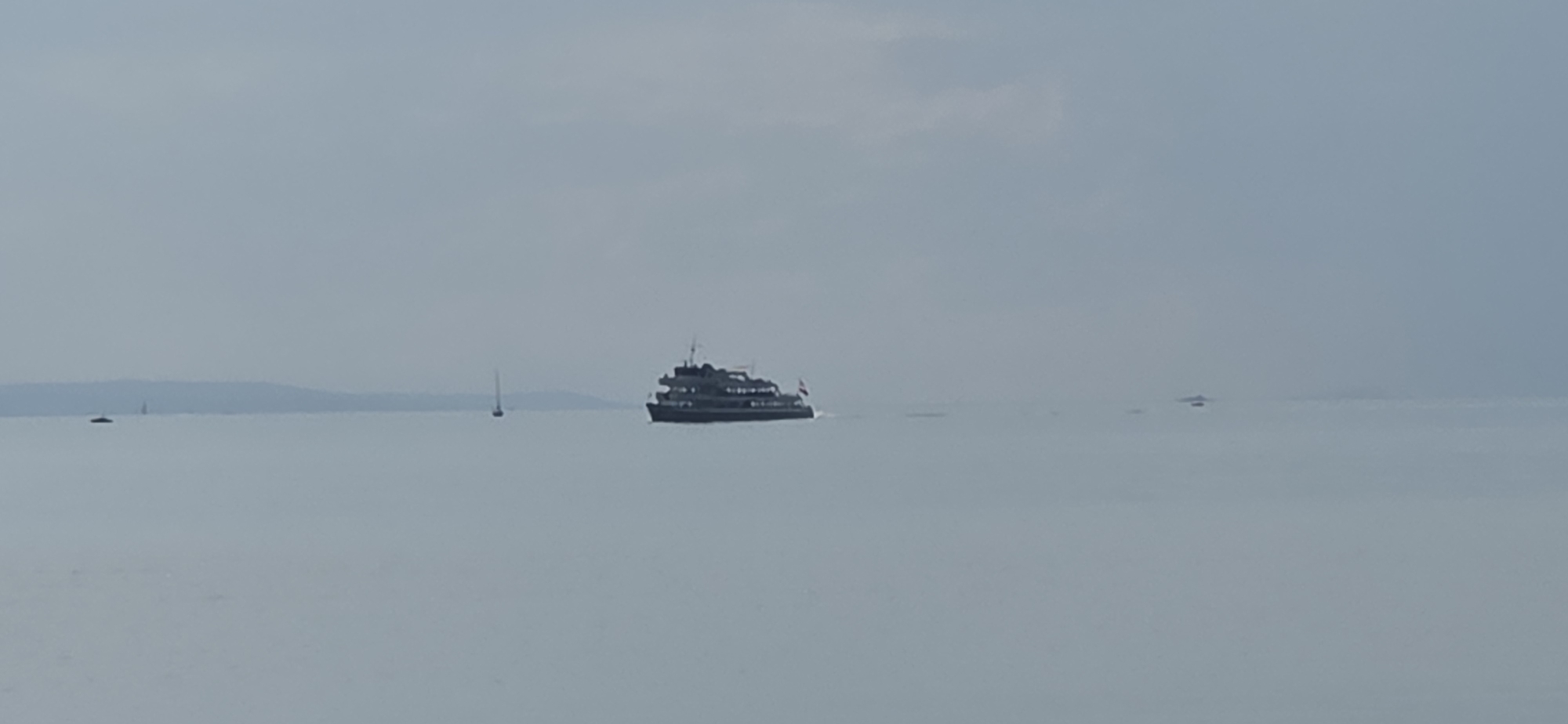 A wide-angle, slightly hazy image captures a calm, overcast day on a body of water, likely a lake given the distant, low-lying landmass on the horizon. The water is a muted, grayish-blue, reflecting the cloudy sky. In the middle ground, a dark, multi-decked passenger boat, possibly a ferry, is seen from its starboard side, moving to the right. It has a modern design with several windows visible on each deck and a flag flying at its stern. To the left of the ferry, a single sailboat with a tall mast is visible, also on the water. Further in the distance, on the horizon, a low, dark landmass, possibly hills or a coastline, stretches across the frame. Several indistinct, darker shapes, likely other boats or structures, are scattered across the water in the far distance. The overall impression is one of peacefulness and openness, with the water and sky dominating the scene.