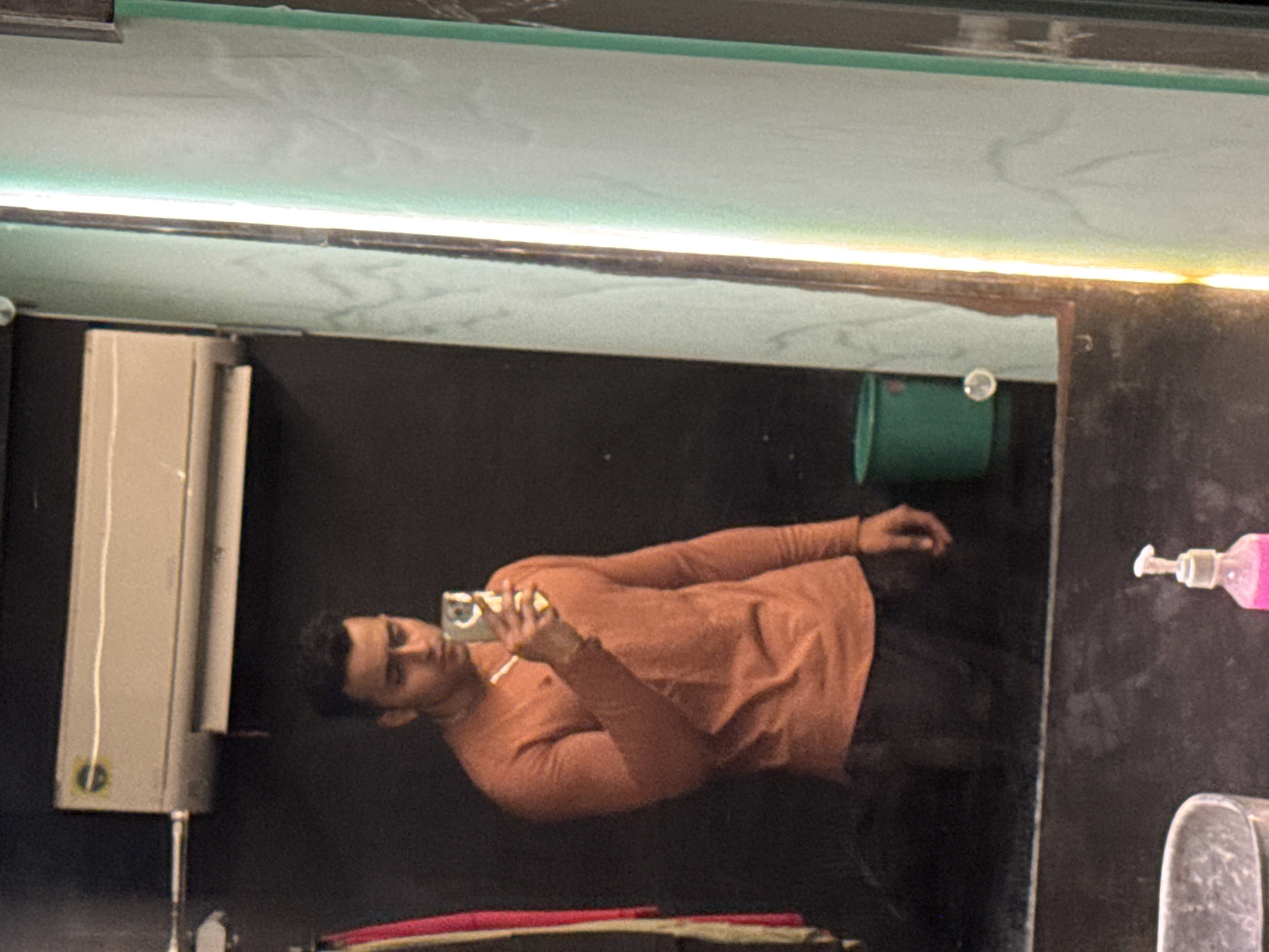 A man is taking a mirror selfie in what appears to be a restroom. He is wearing a long-sleeved orange shirt and dark pants. He is holding a smartphone in his right hand to take the picture. The restroom has a dark, possibly black tiled wall. A white hand dryer is visible on the left. A green bucket is positioned high up, near the top right corner of the mirror's reflection. A soap dispenser and other bathroom items are partially visible to the right. The location might be Kolkata, India.