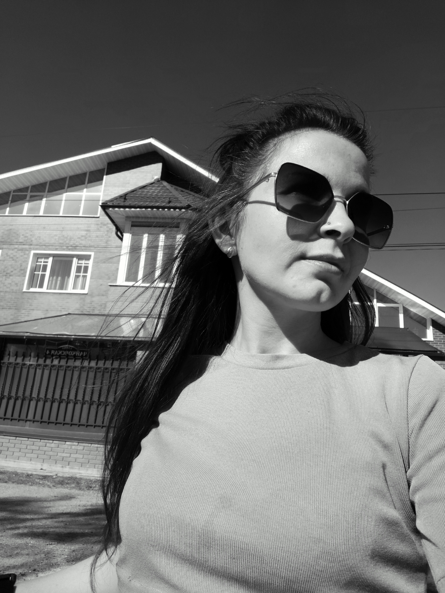 A black and white, close-up shot of a woman wearing oversized sunglasses, with a residential building and fence visible in the background, likely taken in Фоки, Russia.
The woman has dark hair, some strands of which are blowing in the wind. She is wearing a long-sleeved, ribbed shirt. The background includes a two-story brick building with multiple windows, and a dark-colored fence with lettering in Russian. Overhead electrical wires are visible against the sky. The overall setting appears to be a sunny day in a residential area.