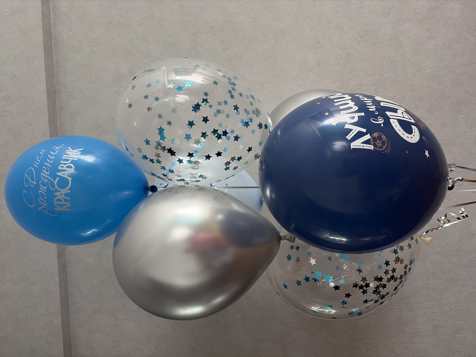 A bouquet of six balloons is displayed against a textured gray wall. The balloons are in various colors and finishes: one is bright blue with white lettering that reads "С Днём рождения, КРАСАВЧИК" (Happy Birthday, Handsome), another is silver and metallic, three are clear with blue star-shaped confetti, and one is dark blue with white lettering and a graphic of a soccer ball. The dark blue balloon has lettering that appears to read "ЛУЧШИЙ в мире сын" (Best son in the world). The balloons are tied together with ribbons and strings. There is no visible indication of the city of Chaikovsky, Russia, within the image itself.