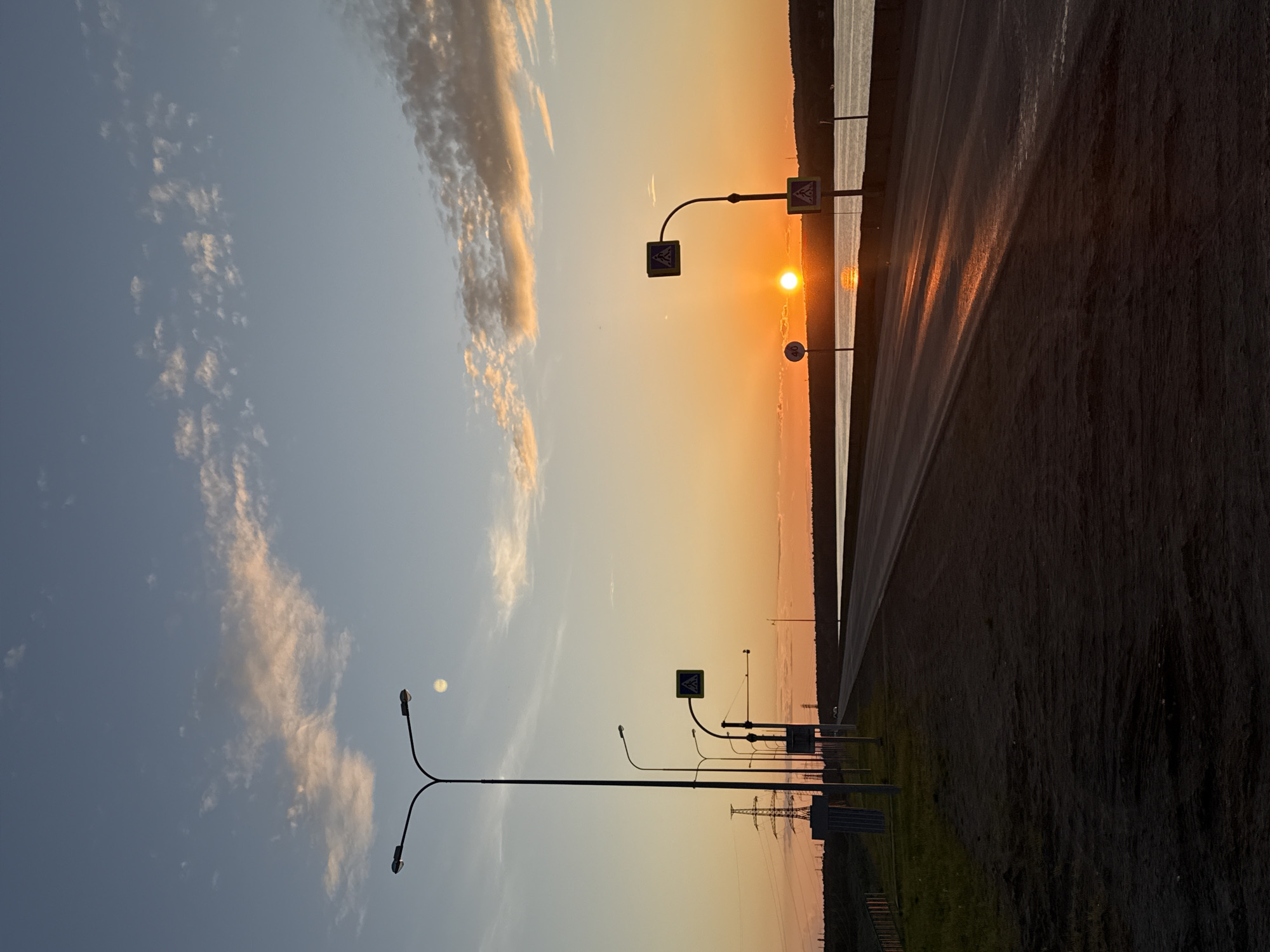 The image depicts an outdoor scene in Chaikovsky, Russia, at either sunrise or sunset, characterized by the warm, low-angle illumination of the golden hour.
The sky transitions from a pale blue with scattered, wispy clouds—some exhibiting light orange coloration—in the upper and middle regions, to a vibrant orange and yellow gradient along the horizon. A distinct, bright orange sun is prominently positioned low on the horizon, above a body of water on the right side of the frame. This sun casts a strong, elongated reflection across the water and onto the adjacent asphalt road surface. High in the upper-left quadrant of the sky, a faint, pale, diffuse disk, consistent with the moon, is visible.
An asphalt road, appearing to be a two-lane thoroughfare, extends from the dark foreground into the distance, running parallel to the body of water. Its surface displays prominent linear reflections from the sun. To the immediate left of the road, a dark, unlit dirt and grassy shoulder gives way to a line of infrastructure. This includes several tall, dark streetlights with double, curved arms, and additional single-arm streetlights receding into the background. A large, dark lattice-style electricity transmission tower is prominent on the left mid-ground, accompanied by power lines. Multiple road signs are present: a blue square pedestrian crossing sign featuring a white triangle and black walking figure is centrally located on a pole, partially silhouetted against the sun. Another identical pedestrian crossing sign is visible further down the road. A circular white speed limit sign with the numeral '40' in black is positioned on the right side of the road, closer to the sun's position.
A wide body of water, consistent with a river or large lake, stretches across the mid-ground, reflecting the intense orange light of the sun and portions of the illuminated sky. Beyond the water, a dark, low-lying landmass, appearing as a treeline or distant hills, forms the horizon. There are no discernible human figures present in the scene. Faint, dark outlines that may be one or two vehicles are barely perceptible in the far distance on the road.