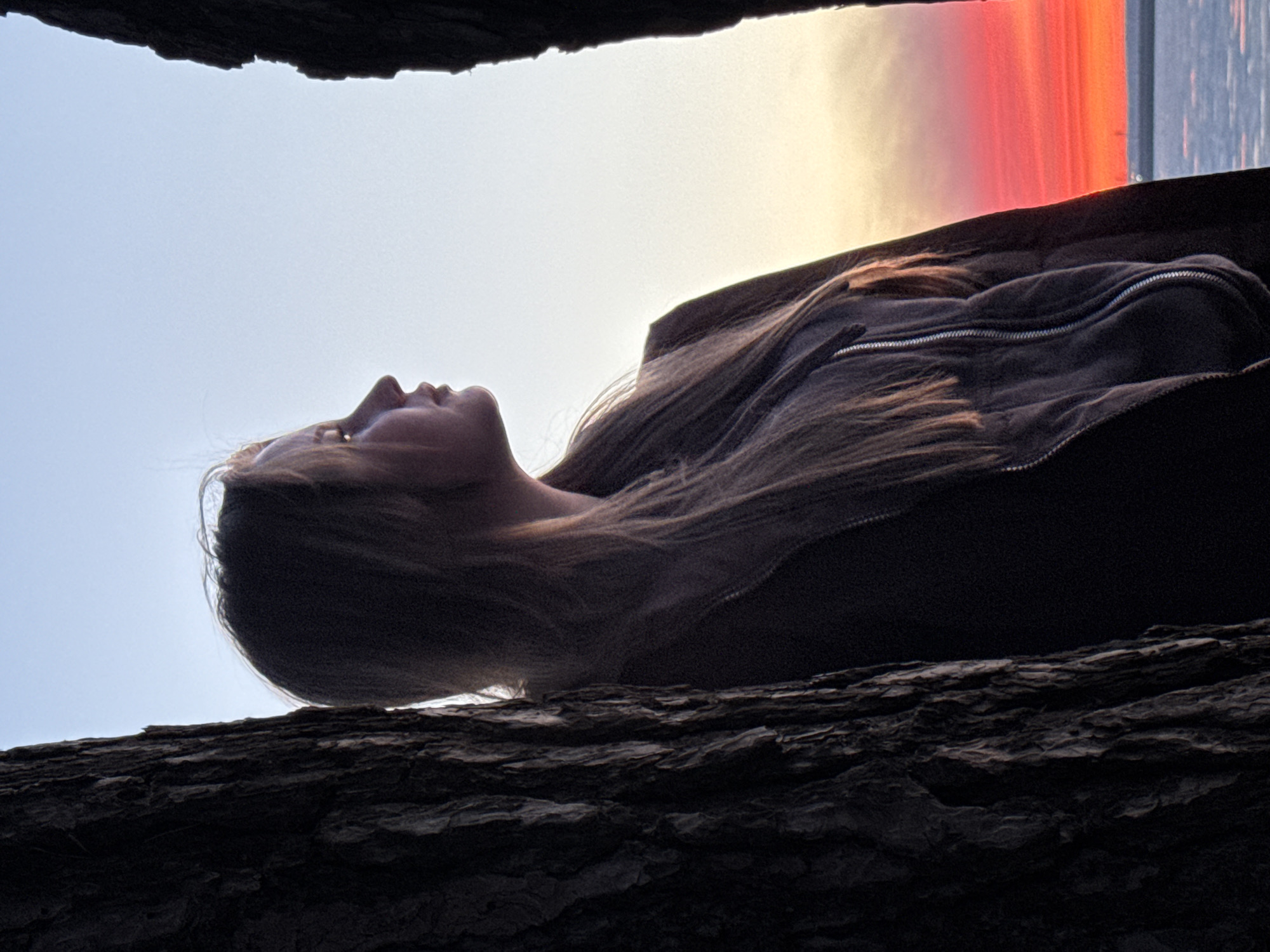 A woman with long blonde hair, wearing a zipped-up sweatshirt, lies on a dark, textured surface (possibly tree bark or rock) with her head tilted back. The backdrop is a sky transitioning from light blue to a vibrant orange and red sunset, likely indicating dusk. Overhanging the scene, at the top, is a dark mass suggestive of rock or a tree branch. The location could be Chaikovsky, Russia.