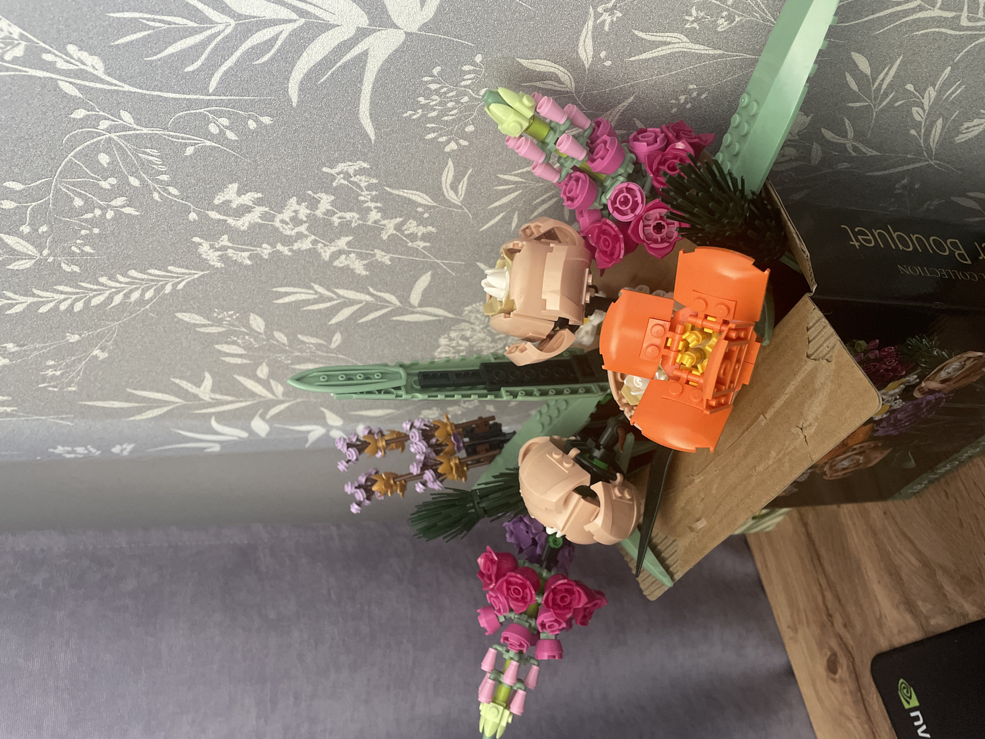 An indoor close-up shot shows a Lego flower bouquet constructed from various colored plastic pieces. The bouquet features pink roses, purple and gold flowers, and green leaves and stems, arranged in a cardboard box. The box is decorated with an image of flowers and text that reads "er Bouquet". To the left of the bouquet, a grey curtain hangs, and behind it, a wall is visible with a subtle floral wallpaper pattern. A wooden desk surface is in the foreground, with a black computer mousepad displaying a green Nvidia logo. The lighting in the scene is neutral, casting soft shadows. The image is located in Unknown, Russia.