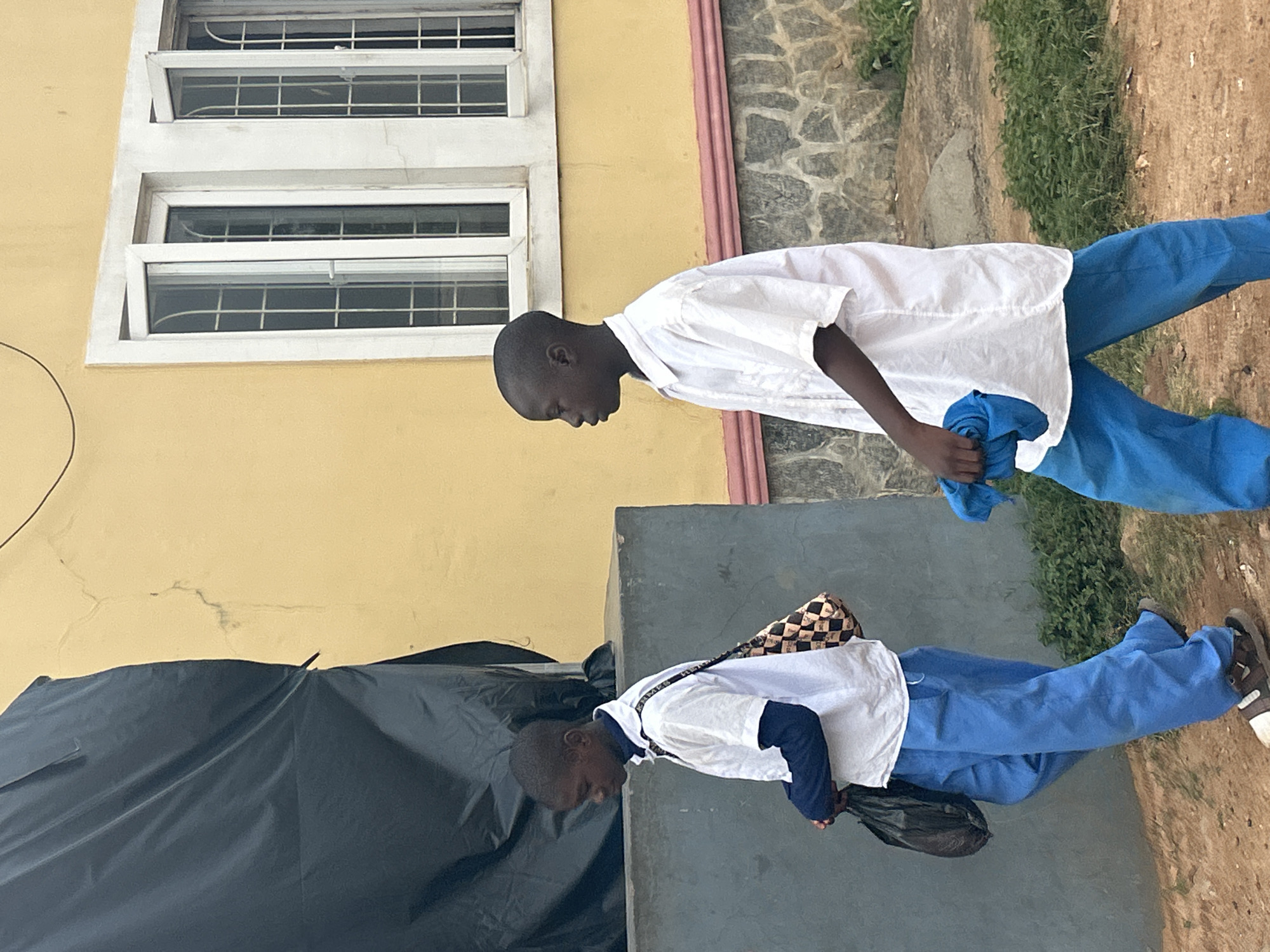 Two Black male children, presumably schoolchildren given their uniforms of blue trousers and white shirts, are pictured outdoors near a building. The building is pale yellow with white-framed windows. The ground is mostly dirt with some grass and a stone or concrete wall. The location is likely Bar Jirgi Summa, Nigeria. One child is carrying a blue cloth, the other a black bag and checkered satchel. The context surrounding them and their actions are undefined.
