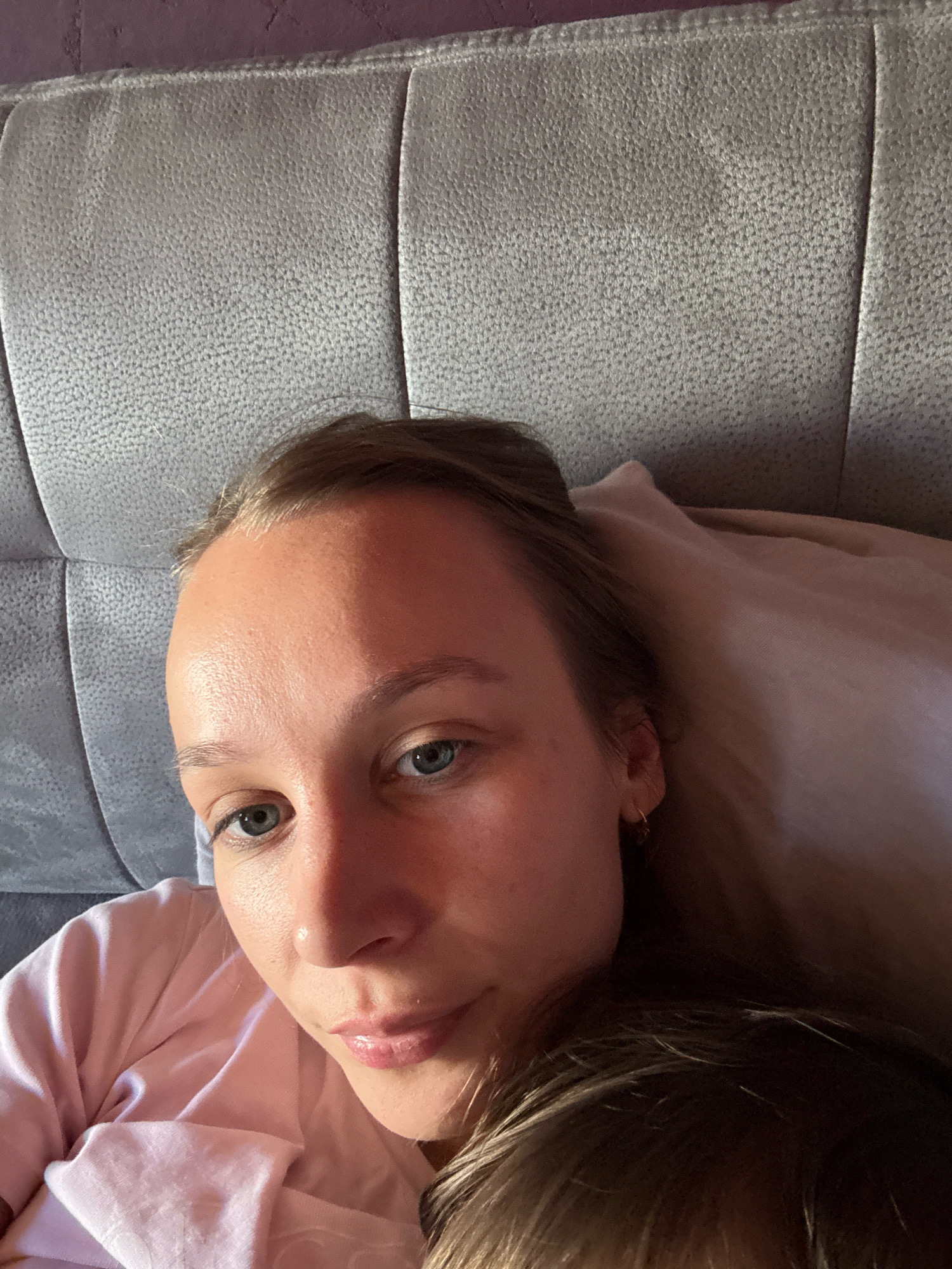 The image is a close-up, high-angle selfie of a young woman lying down. She has fair skin, blue eyes, and light brown hair. She is wearing a pink shirt and a gold hoop earring. The background consists of a textured, light gray headboard and a purple wall. The woman's face is partially illuminated by sunlight. There is no visible indication of the location being in Überherrn, Germany.
