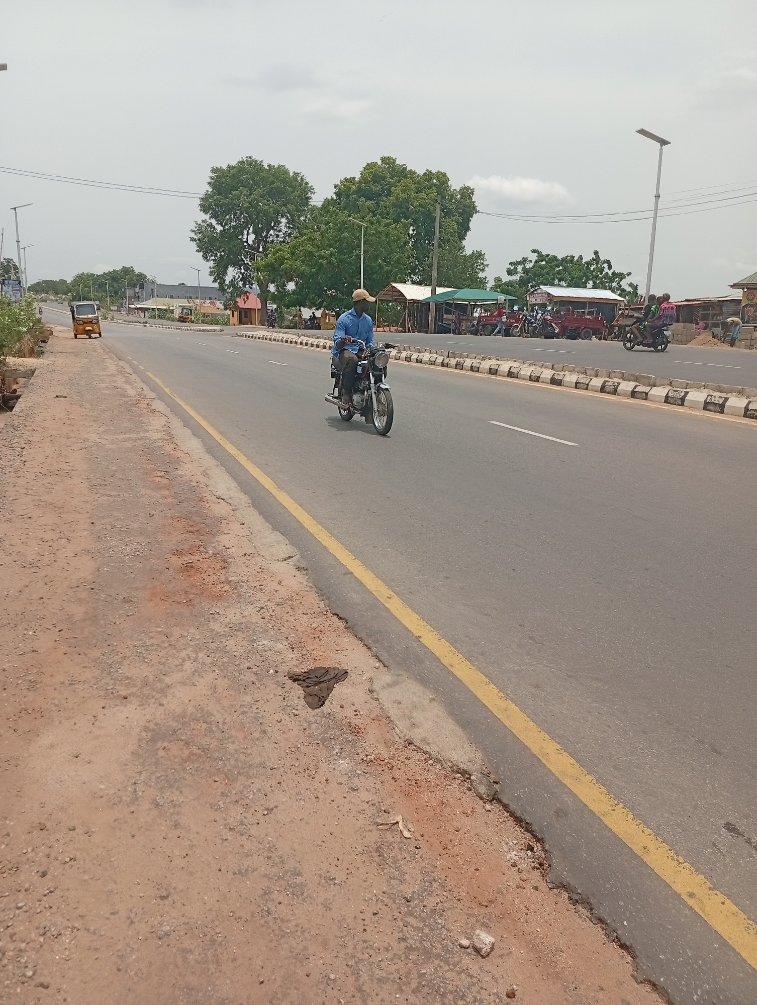 The image depicts a wide paved road in Adankolo, Nigeria. A motorcyclist in a blue shirt and cap is riding in the center lane. A yellow line marks the edge of the road, with a dirt shoulder to the left. The road has a center divider with black and white blocks. In the background, there are trees, buildings, and people gathered around what appears to be a marketplace. Other vehicles, including a tricycle, are visible on the road. Streetlights line the side of the road. The sky is overcast.