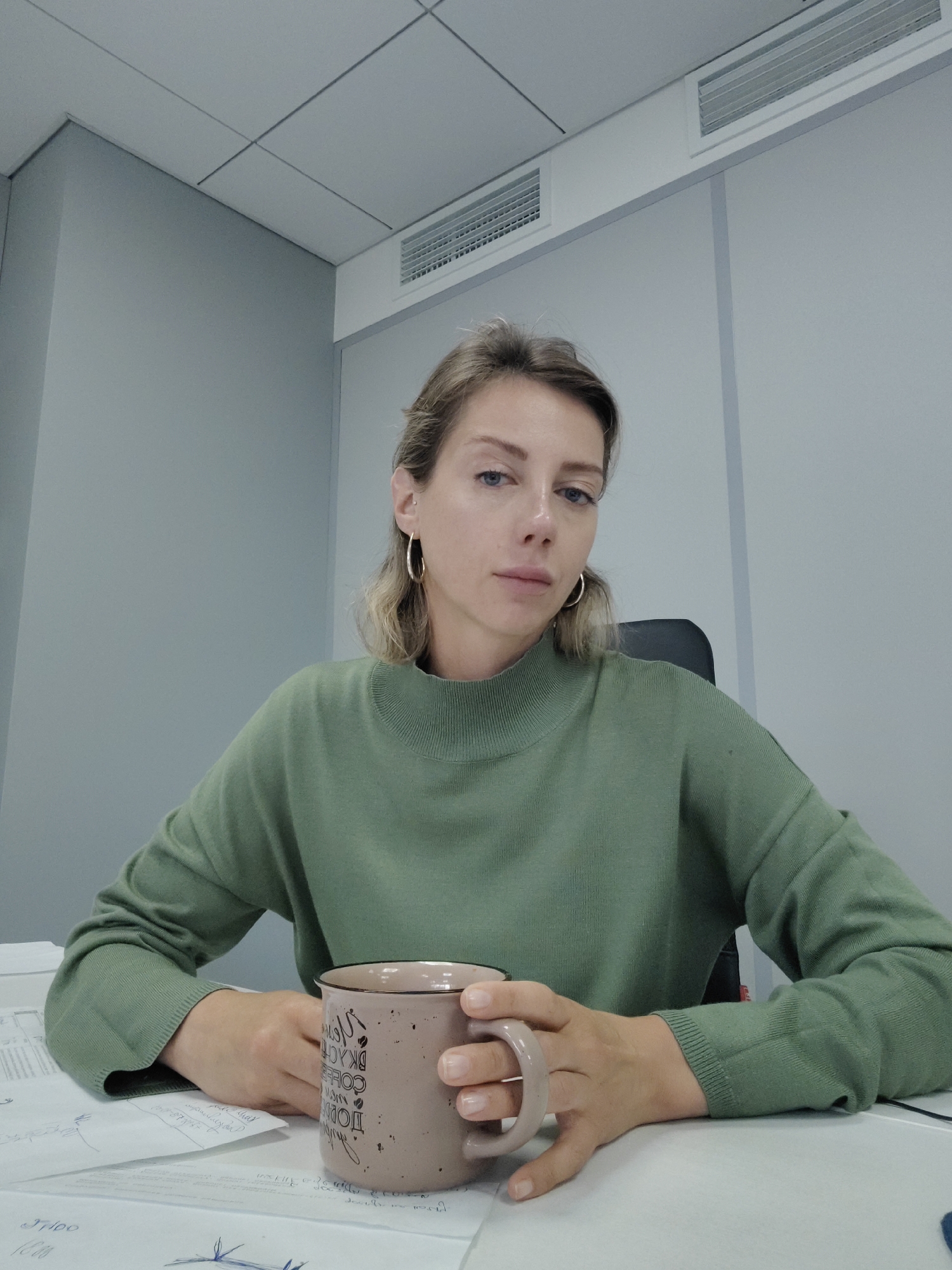 A Caucasian woman with blonde hair pulled back, wearing a green turtleneck sweater and gold hoop earrings, is seated at a desk with papers and a mug. She is holding the mug with both hands. The background appears to be an office with grey walls and a white tiled ceiling with ventilation grilles. The scene is indoors and well-lit. The location is not specified in the image or the provided crops.