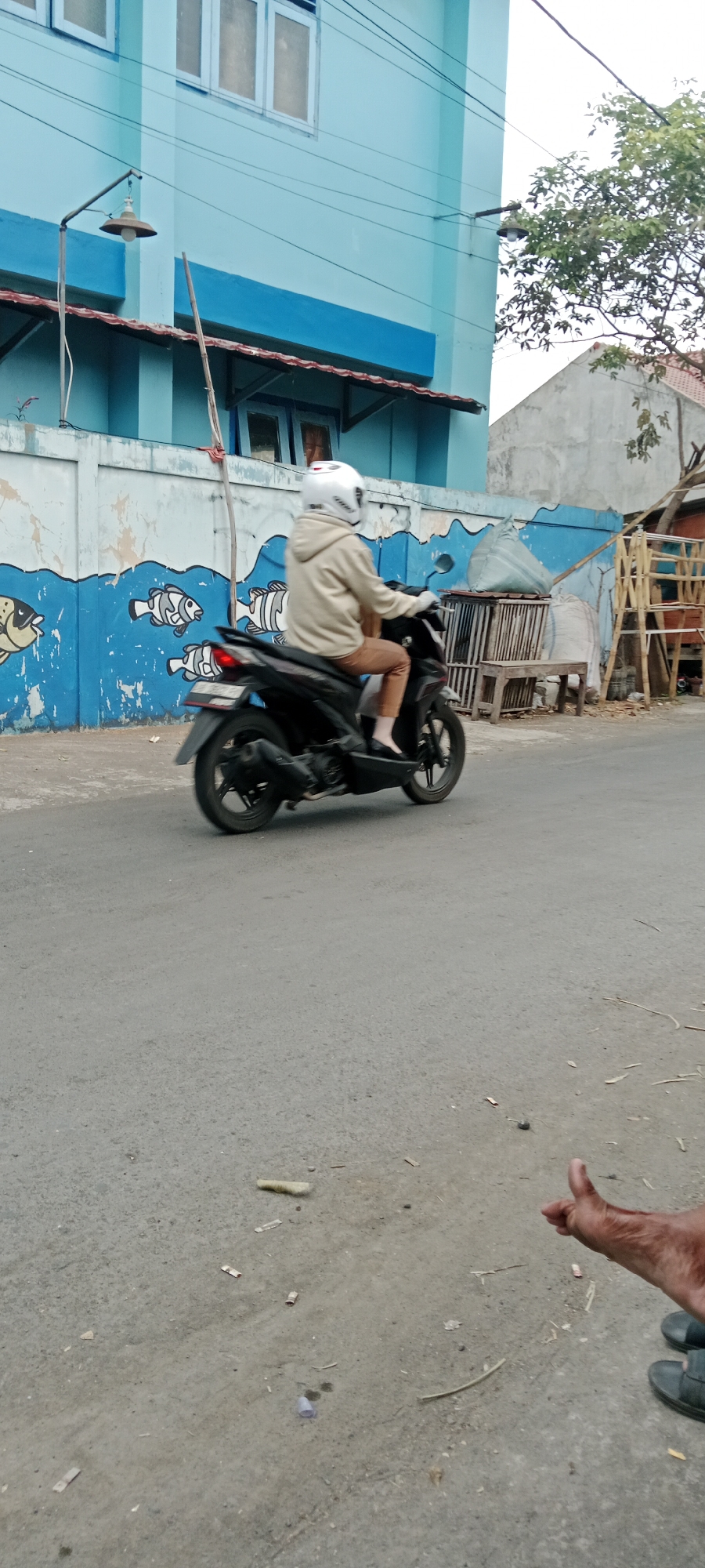 A person wearing a white helmet, beige hoodie, and brown pants rides a black scooter on a paved road in Purwomartani, Indonesia. The scooter is in motion, indicated by the blur of the wheels. To the left of the road, a blue building with white trim and windows is visible. Below the building is a low wall adorned with a mural of blue waves and white fish. To the right of the scooter, a wooden structure with stacked materials and a wooden bench is present, with a large tree branch extending over the scene. The road surface is asphalt with scattered debris, including small pieces of paper and what appears to be a plastic cup. In the foreground, a person's hand is visible, giving a thumbs-up gesture. The overall scene depicts a street view in a residential or commercial area of the town.