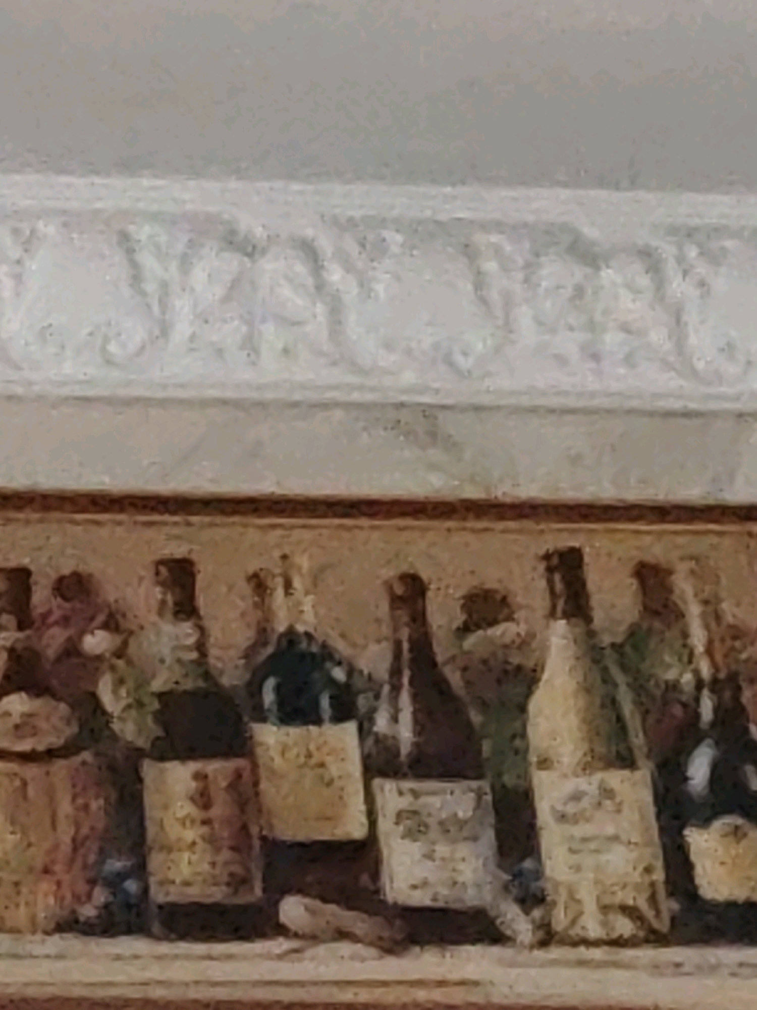 The image depicts a still life painting, likely a decorative piece, featuring a collection of bottles and possibly other decorative objects arranged on a shelf or ledge. Above the objects is a decorative white molding, likely part of a wall or furniture. The overall style suggests an artistic rendering rather than a photograph. The painting is of moderate quality with visible brushstrokes and a slightly aged appearance. There are no people or active interactions present in the scene. The background is a muted, light-colored wall. The specific location in Ninotsminda, Georgia, is not discernible from the visual information provided in the image.