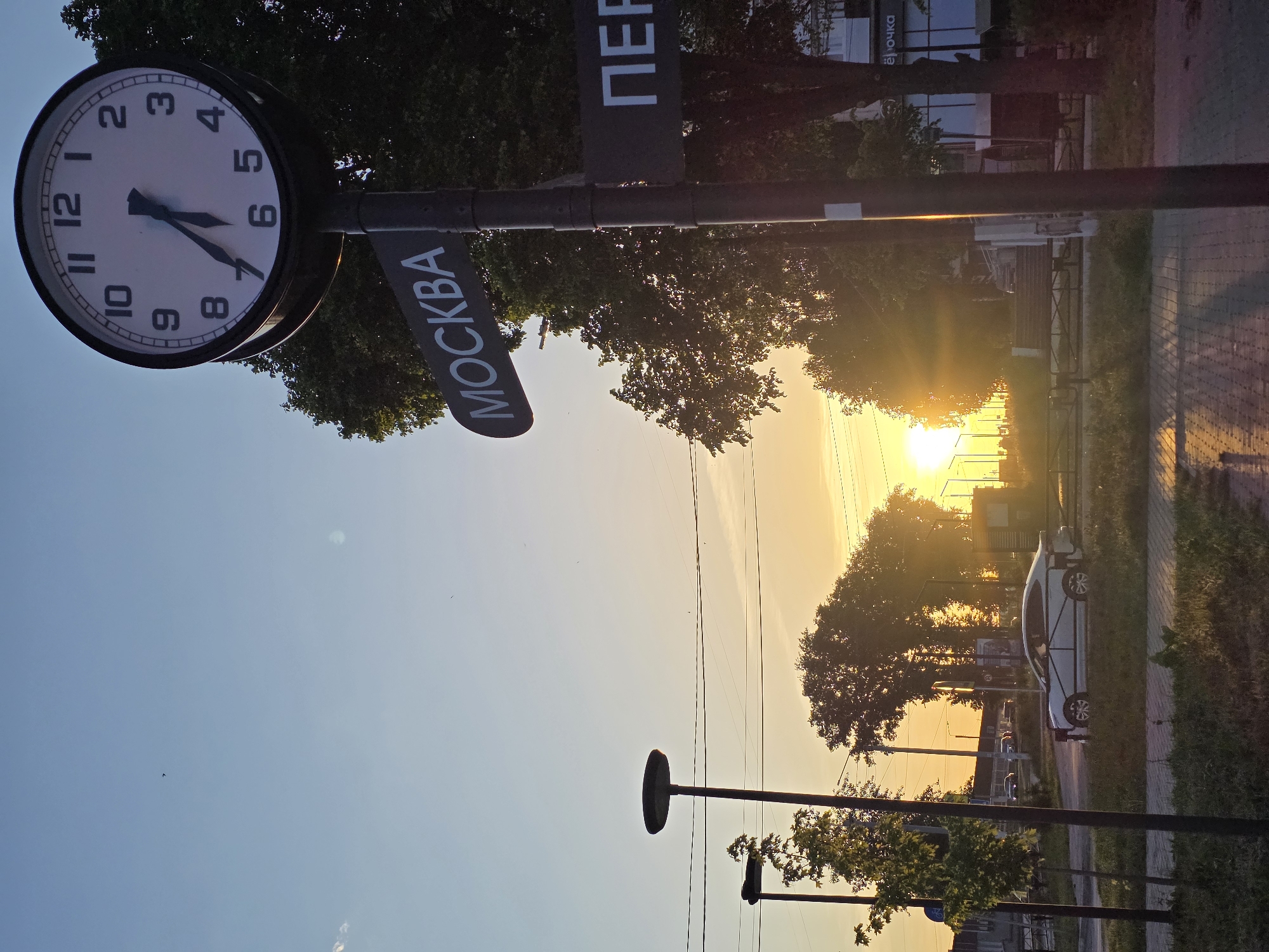 The image depicts a street scene in Chaikovsky, Russia, at sunset. A large, circular clock is mounted on a pole, displaying approximately 5:35. Below the clock are signs in Russian, with "Москва" (Moscow) visible on one. "Пер" is visible on another. A sidewalk runs along the right side of the frame, with parked cars and buildings visible in the background. Trees line the street, silhouetted against the setting sun. Power lines stretch across the sky. A streetlight is also visible.