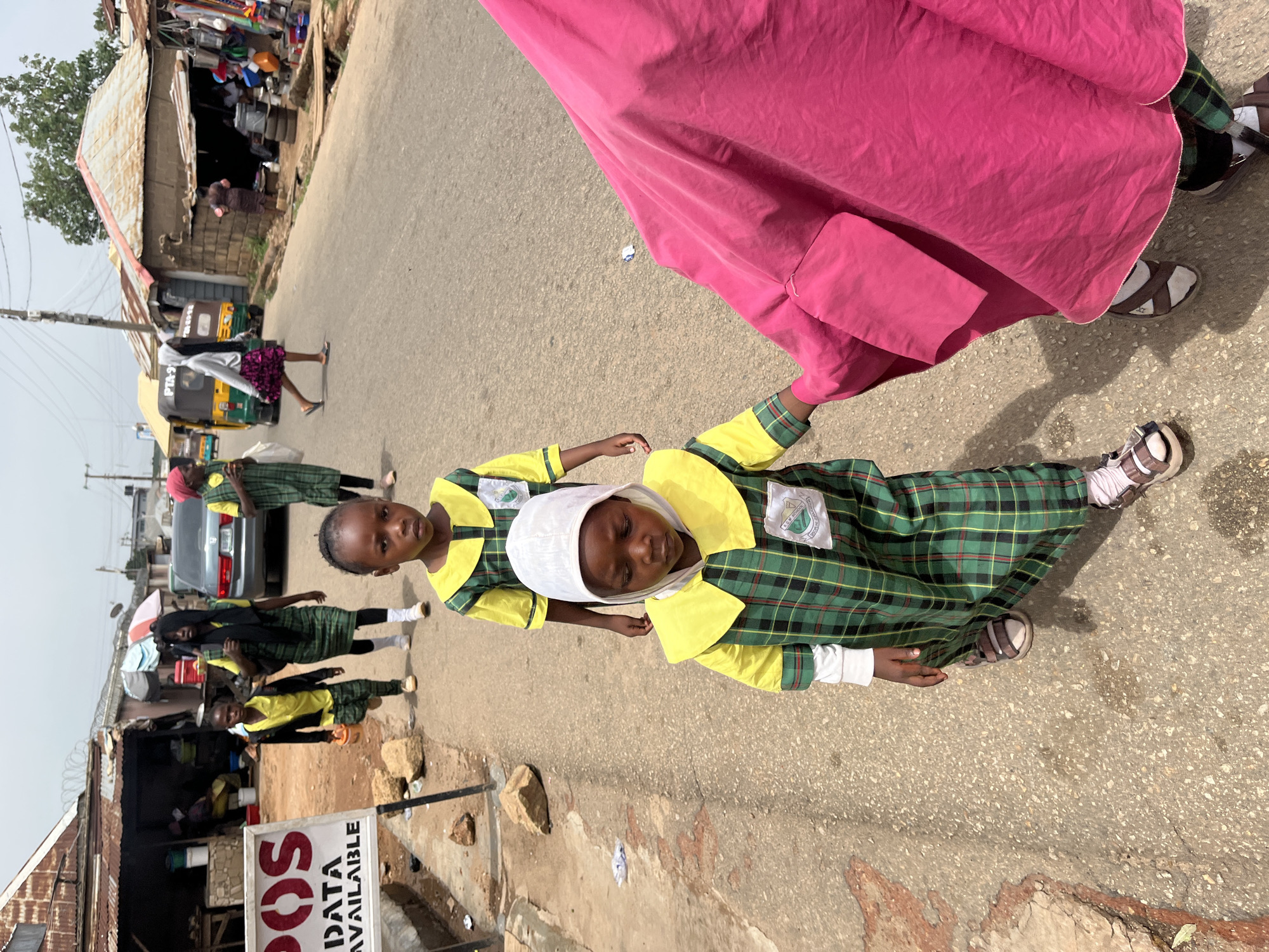 The image shows two girls in green and yellow plaid school uniforms in what appears to be a street in Jos, Nigeria. One girl is helping the other, who is wearing a white head covering and lying down on the asphalt road. A person in a long, pink robe stands nearby. In the background, there are buildings, parked vehicles, and other people walking on the side of the road. A sign reading "POS Data Available" is visible near the foreground. The overall scene suggests an urban or semi-urban area with commercial activity along the roadside.