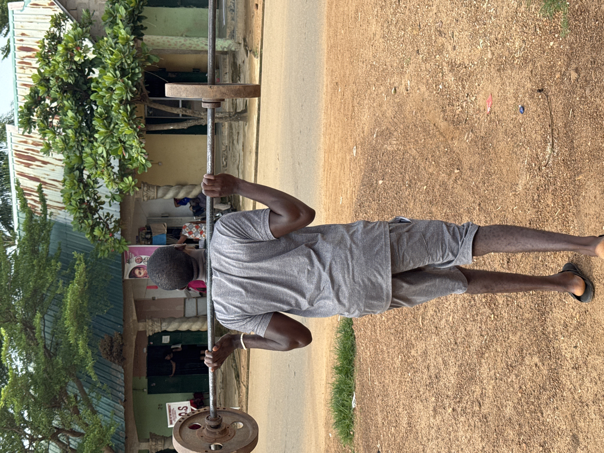 A young Black male in Akwanga, Nigeria, stands outdoors, holding a barbell on his shoulders. He is wearing a grey t-shirt and grey shorts, and flip-flops. The barbell has two large circular weights attached to each end. The background shows a street with buildings, some with corrugated metal roofs and others with painted facades. Trees and vegetation are visible, as are other people in the distance. The ground is covered in reddish-brown dirt and gravel. The scene appears to be a residential or commercial street.