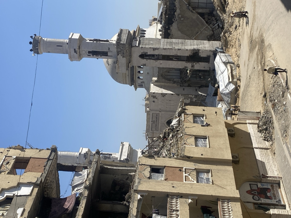 The image depicts a devastated urban landscape in Khan Yunis, Palestinian Territory, under a clear blue sky. On the left, a multi-story building, appearing residential or commercial, exhibits severe structural damage, with upper floors partially collapsed, exposed interiors, and shattered or covered windows. A sign on the ground floor reads "YASEEN GAMES" with a PlayStation logo. Piles of rubble lie at its base and on damaged sections. In the midground, a prominent mosque with a beige dome and a tall, white minaret topped with loudspeakers shows significant damage to its walls and exterior, though its dome appears mostly intact. Behind the mosque, another damaged building with an arched facade and an Arabic inscription is visible.
The ground between these structures is an unpaved dirt road strewn with debris. Several collapsed or damaged white tarpaulin shelters are visible in the midground. Three distinct groups of people are present: one individual walking away on the foreground road, carrying a dark bag; a small group of at least three individuals gathered near the shelters; and two individuals walking on a path in the background to the right. A single black wire spans horizontally across the upper part of the frame. The scene is illuminated by bright daylight.