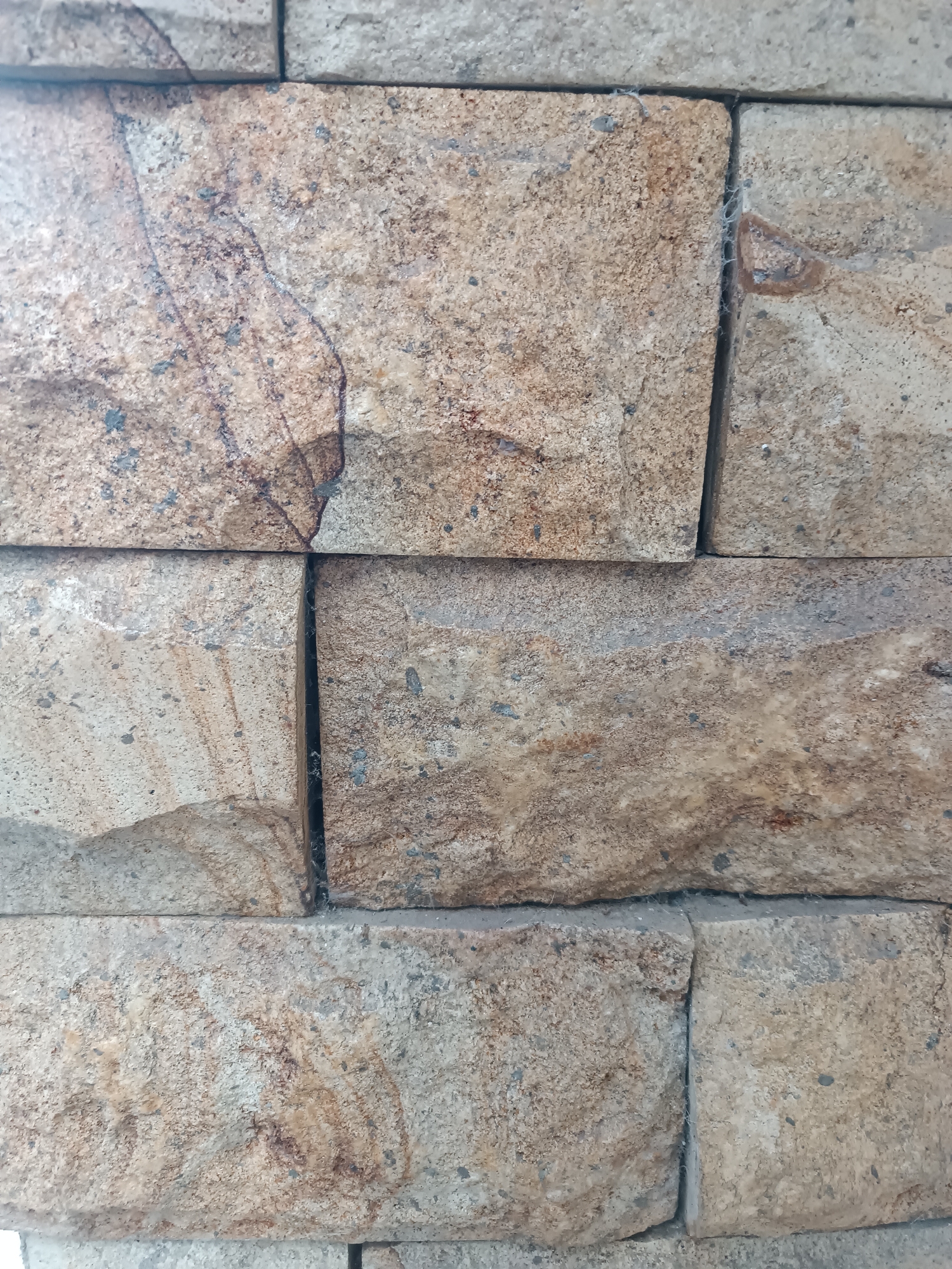 The image displays a close-up view of a wall constructed from rectangular stone tiles. The tiles are predominantly light brown and beige, with visible variations in texture and color, including some darker brown and gray mineral deposits and veins. The stones are arranged in horizontal rows with slight offsets, creating a stacked appearance. The grout lines between the stones are visible and appear to be a lighter gray color. Some of the stones exhibit rough, natural surfaces with visible grain and small imperfections. There are also indications of very fine cobwebs in the crevices between some of the tiles, suggesting the wall may be outdoors or in an undisturbed area. No people or specific environmental context beyond the wall material itself are discernible. The location in Tabanan, Indonesia, is not visually represented in the image.