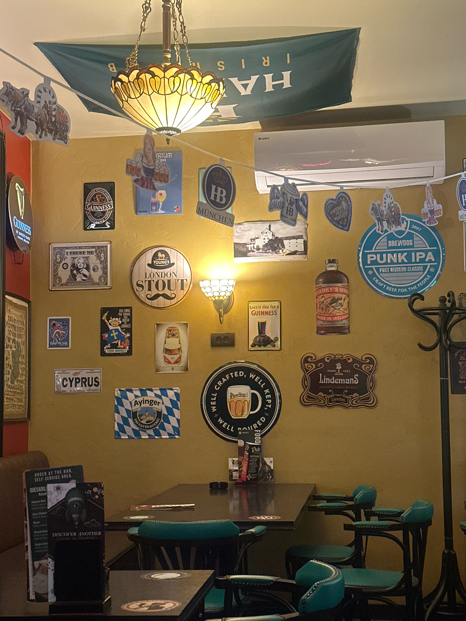 The image depicts the interior of a bar or pub. The walls are painted a warm yellow and are adorned with numerous decorative signs and posters related to beer brands and local establishments. A large green banner with white lettering, possibly "HARRIS," hangs from the ceiling. A decorative stained-glass chandelier is also visible. The scene includes tables and chairs, some of which are upholstered in teal. There is a coat rack on the right side of the image. The overall atmosphere suggests a casual and somewhat vintage setting. The location is identified as Belgrade, Serbia.