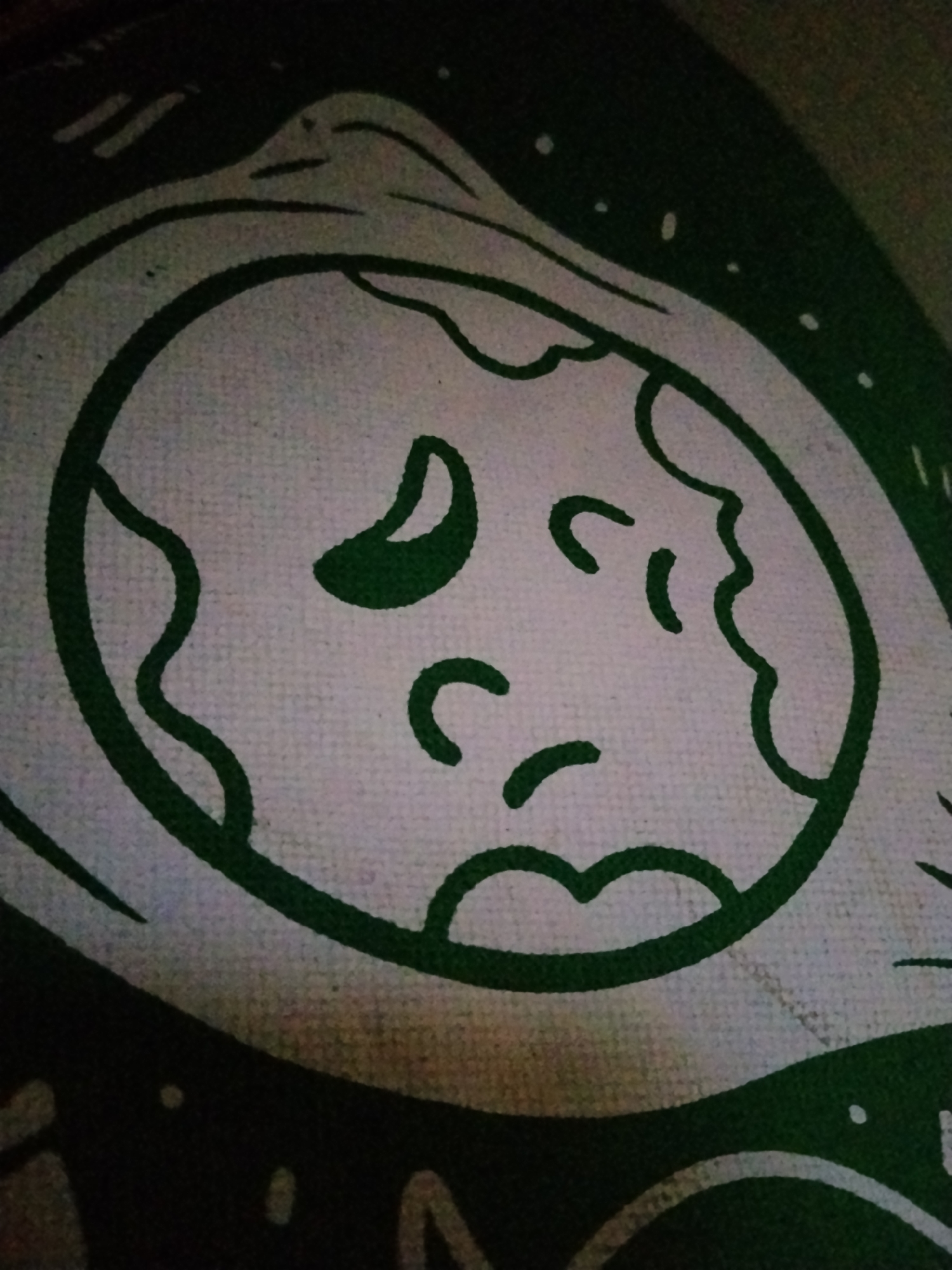 The image shows a close-up of a green and white design on a textured surface, likely fabric. The design features a stylized face with large, dark green eyes and curved green lines representing eyebrows, a nose, and a mouth. The face is enclosed within a larger, uneven green circle, which is part of a more complex pattern that includes abstract shapes and dots. The lighting is dim, and the angle of the shot suggests the object is lying flat. There is no indication of a specific city or location.