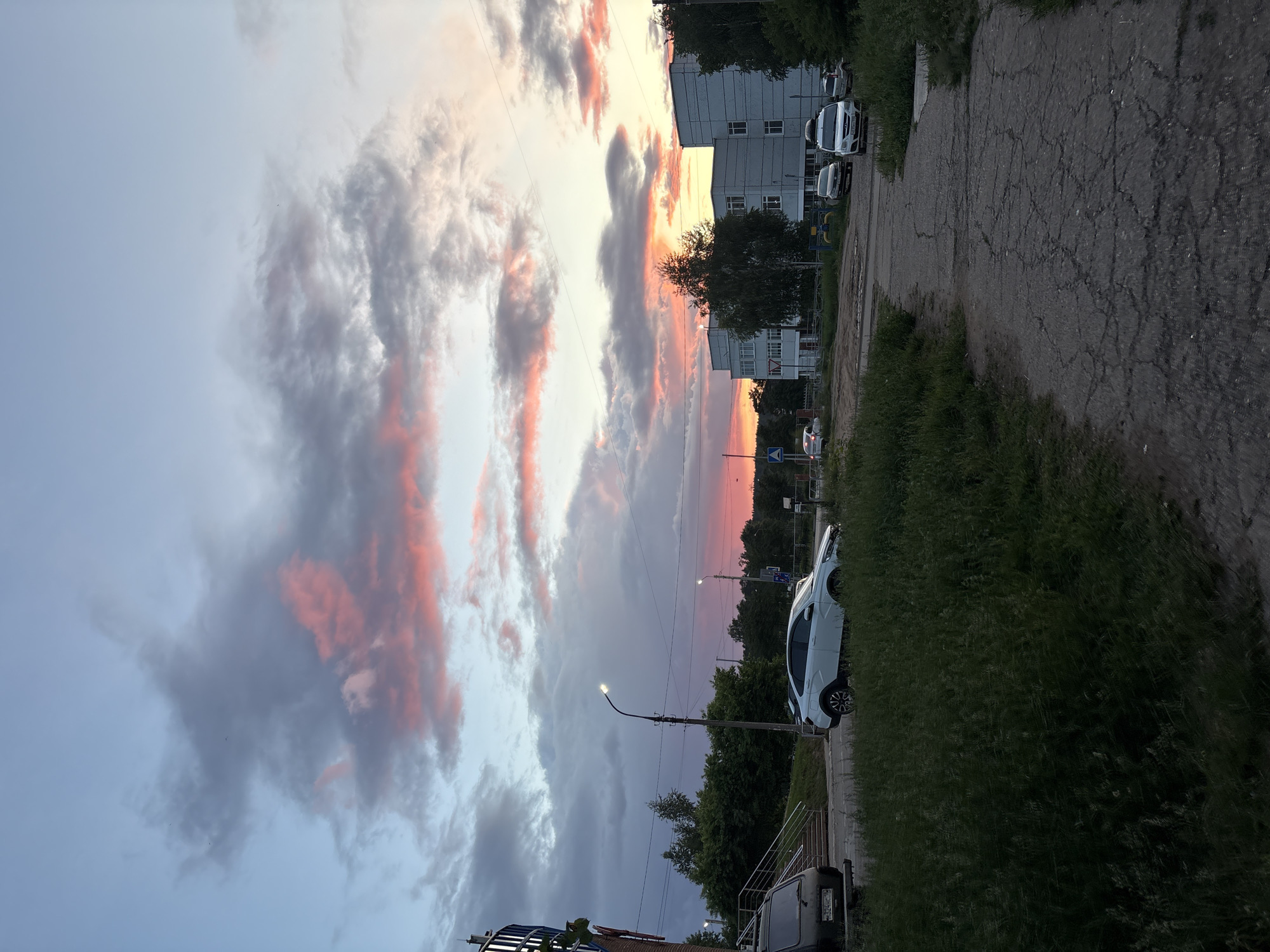 The image depicts a street in Chaikovsky, Russia at dusk. The sky is filled with clouds tinged with pink and grey, suggesting a sunset. A multi-story building is visible along the right side of the frame, with several parked cars in front. The road is asphalt with visible cracks and damage. Grass and vegetation line the side of the road. A street lamp is visible on the right, partially obscuring the pink sky behind. Other street features include trees, road signs, and other vehicles. The overall scene conveys a quiet urban environment at the end of the day.