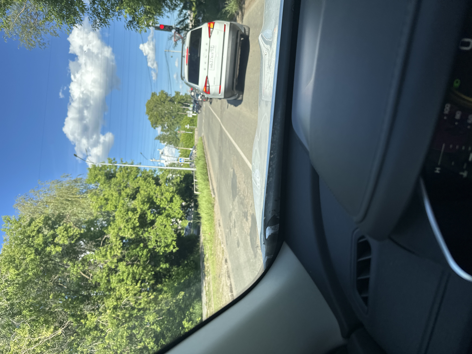 A view from inside a car, with a white Škoda vehicle in front, stopped at a red traffic light. Lush green trees line the road on the left, and a vibrant blue sky with fluffy white clouds is visible above. The dashboard of the car in the foreground is gray and partially obscures the right side of the view. The location appears to be a city street in Unknown, Unknown.