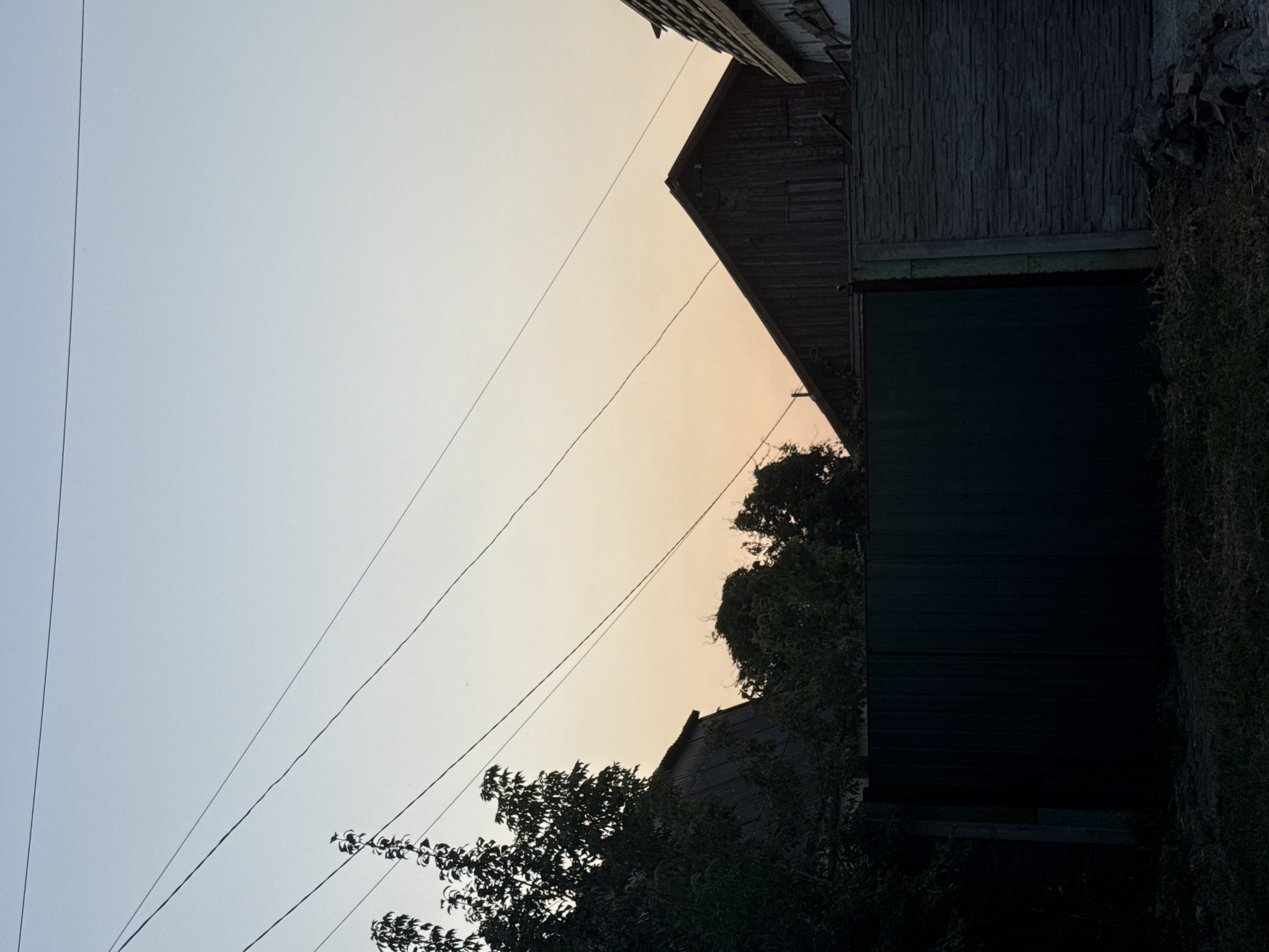 The image depicts a rural or suburban scene at dusk. The sky is a gradient of pale blue and a soft orange, suggesting the sun has recently set or is about to rise. Several power lines traverse the sky diagonally. In the foreground, a dark green metal fence runs horizontally across the frame, partially obscured by dark foliage on the left. To the right of the metal fence, a section of a grey stone-patterned fence is visible. Behind the fences, the roofs and upper portions of wooden structures, likely houses or outbuildings, are visible. The overall atmosphere is quiet and still. The location is identified as Zaporizhzhia, Ukraine.
