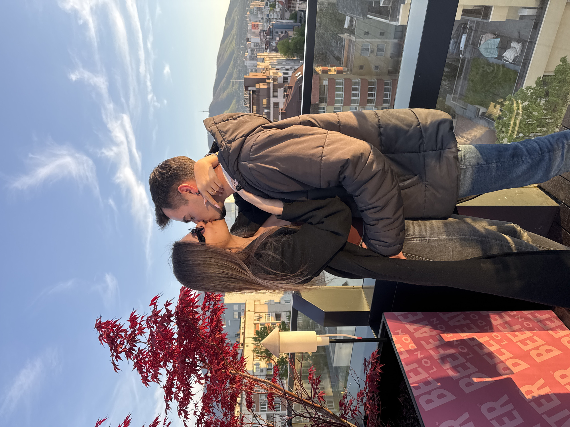The image depicts a couple kissing on a rooftop in Sofia, Bulgaria. The man is wearing a black puffer jacket and jeans. The woman is wearing a black top and sunglasses. They are standing near a glass railing, with a view of the city and mountains in the background. In the foreground, there is a red Japanese maple tree and a table with "Beefeater London" written on it.