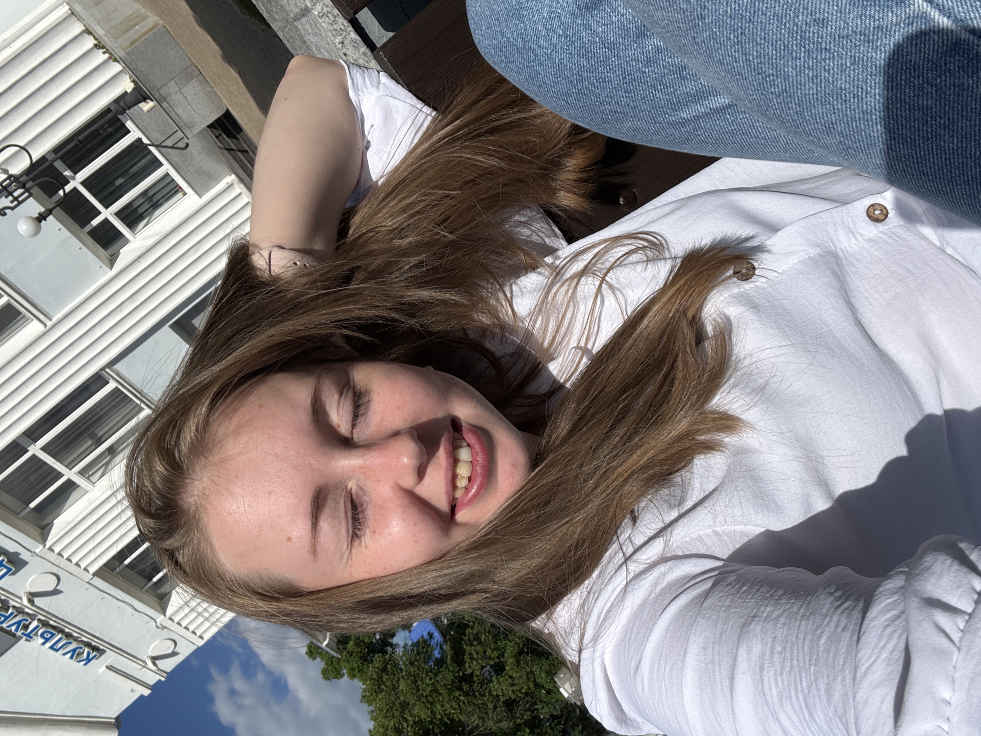 A young adult female with long, light brown hair is depicted in a close-up, outdoor setting. She is smiling with closed eyes, displaying her upper teeth which appear somewhat yellowed, and has visible freckles on her face. She is wearing a white collared shirt with visible brown buttons and blue denim jeans. Her left arm is bent, and her hand rests behind her head or neck, on a dark brown bench or similar surface. In the background, a white building with vertical fluted elements and windows is visible at an angle. Blue Cyrillic text, reading "КУЛЬТУРЫ" (KULTURY), is affixed to the building's facade. An ornate black street lamp with a white globe light stands in the upper right background. Green foliage from trees is visible in the lower left background, beneath a blue sky with scattered white clouds. The scene is brightly illuminated by direct sunlight, casting distinct shadows on the woman's clothing and under her chin. The image is taken in the city of Unknown, Unknown.
