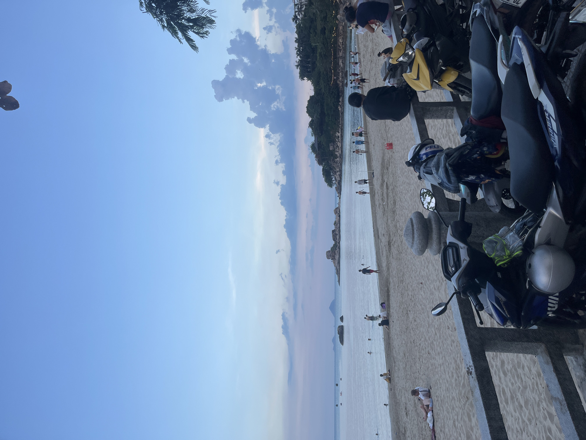 The image shows a beach in Bac Nha Trang Ward, Vietnam. The view is partially obscured by parked motorbikes and scooters in the foreground. A concrete barrier separates the parking area from the beach. People are visible on the sand and in the water. A strip of land with trees and buildings is present in the distance. The sky is blue with some clouds.