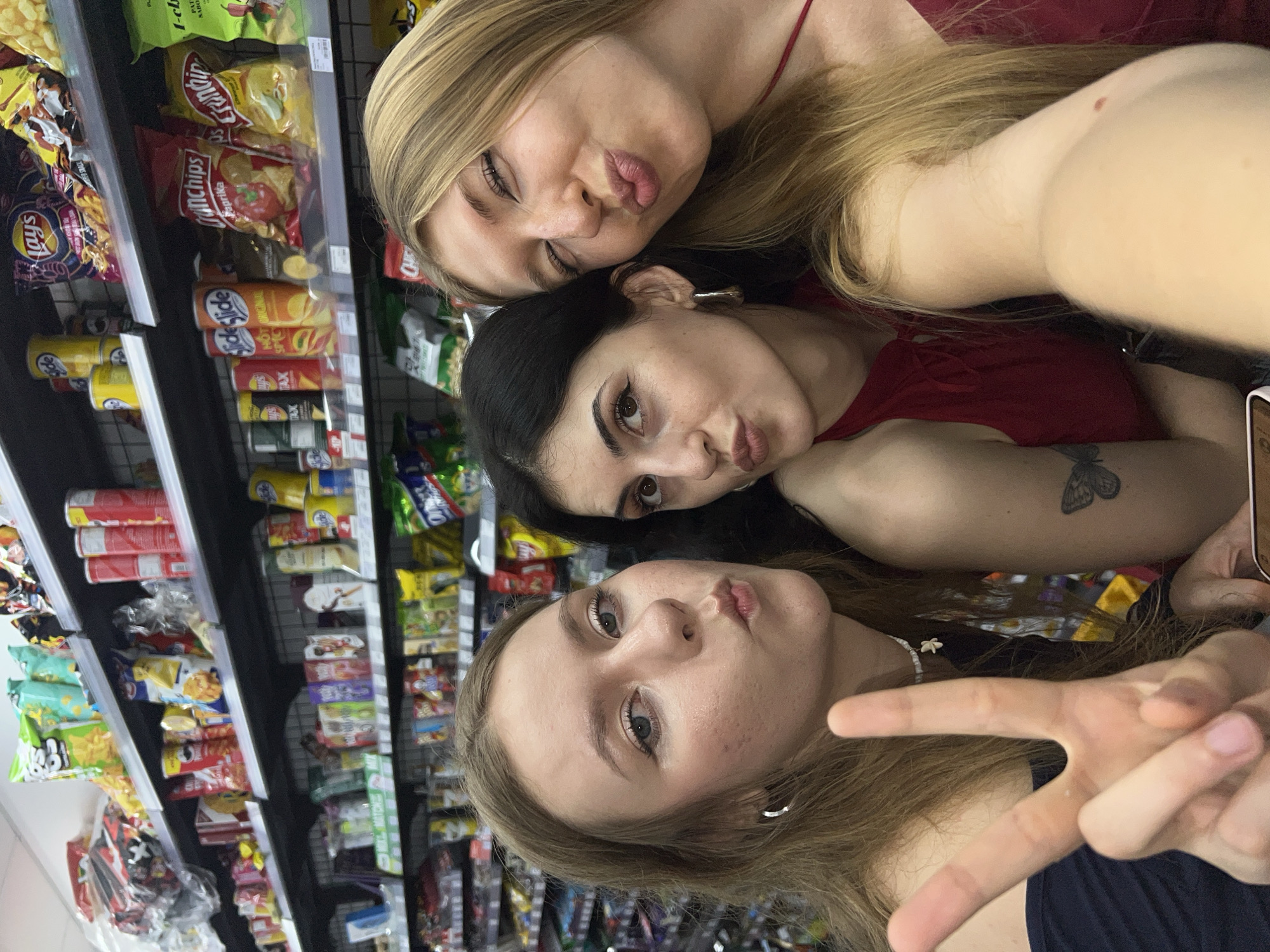 Here is a description of the image:
Three young women are taking a selfie in a store aisle. The aisle is filled with snacks like chips and other packaged goods on shelves behind them. The woman in the bottom center of the frame is blonde with long hair and wearing a dark shirt. She is making a peace sign with her fingers. Above her is a woman with dark hair wearing a red top. Above her is another blonde woman wearing red. All three are making a "kissing" face toward the camera.