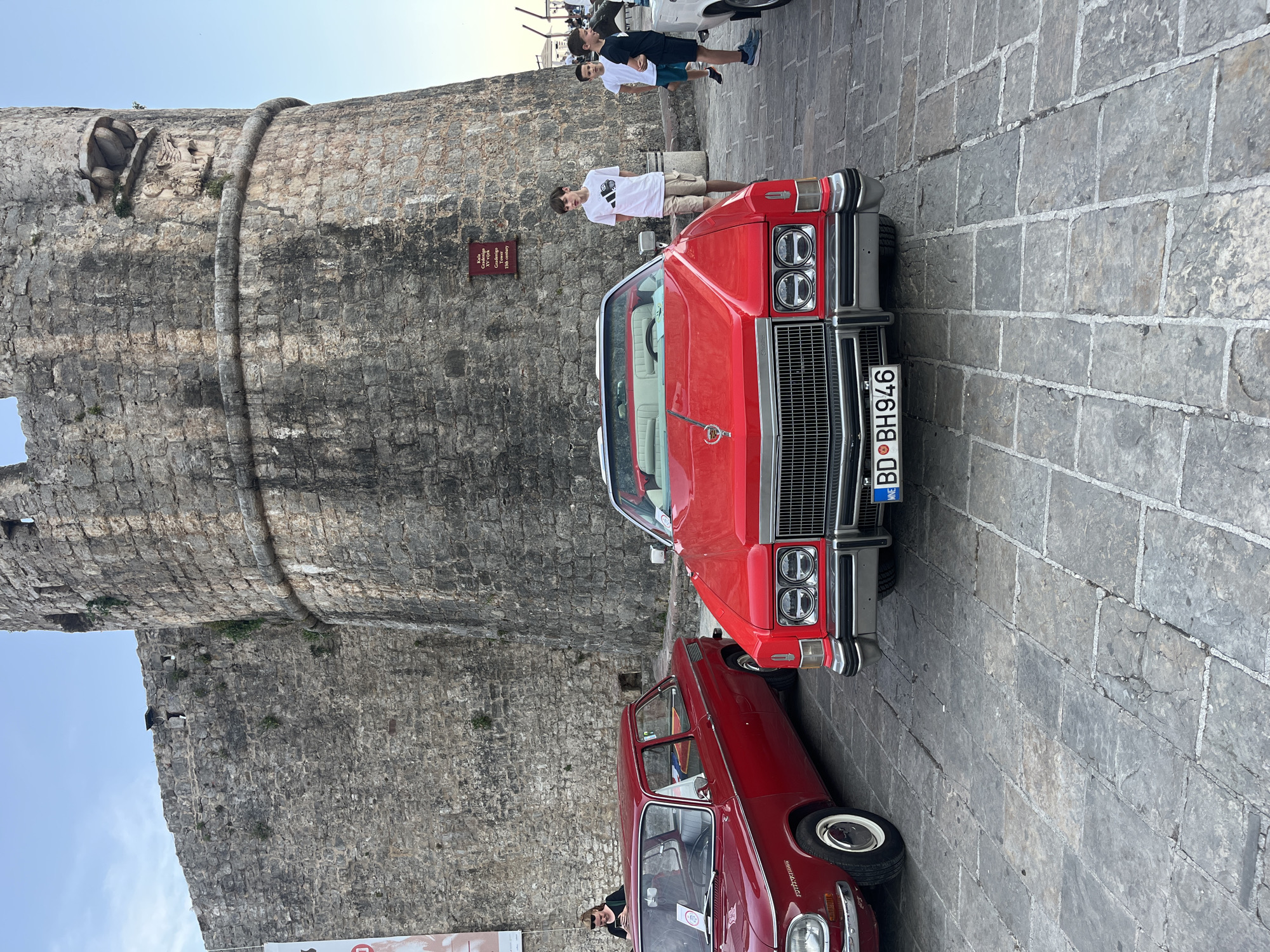 The image shows a view of the old city of Budva, Montenegro. In the foreground, there are two vintage red cars parked on the cobblestone street. The larger car is a convertible and has a visible license plate with the letters "BD BH 946". People are seen in the vicinity of the cars. In the background, there is a massive stone tower forming part of the old city walls. A small sign is affixed to the wall. A few people are standing near the tower and the cars. The sky is light blue. The scene appears to be a tourist attraction or a vintage car show.