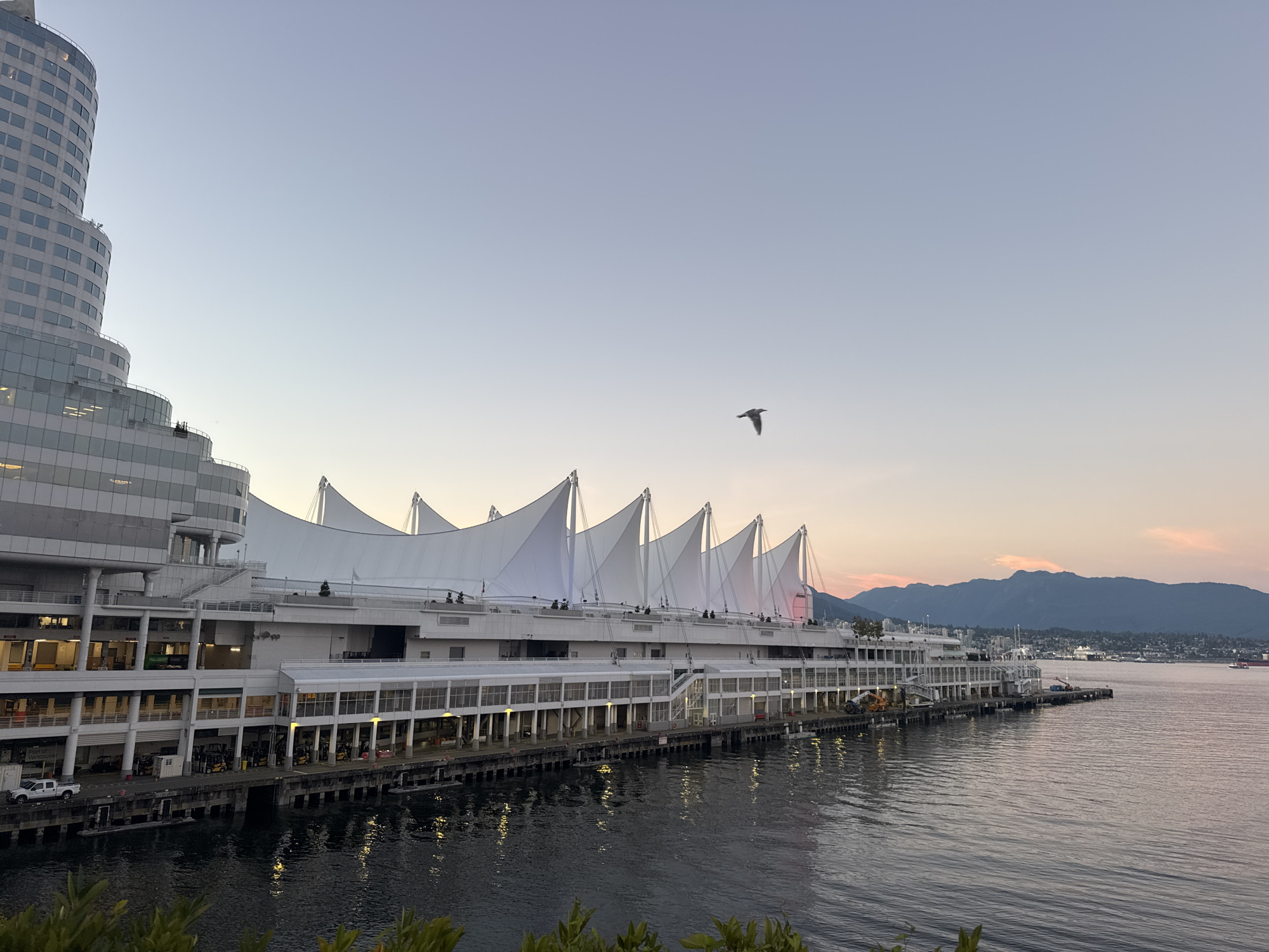 The image depicts Canada Place in Vancouver, Canada, at dusk. The iconic building, known for its sail-like roof structures, is illuminated and reflects on the calm water of the harbor. The sky displays a gradient of soft blues and pinks, indicative of the sunset. In the foreground, a bird is captured mid-flight against the vast sky. The water is dark and still, with reflections of the building's lights visible. Beyond Canada Place, a mountain range forms the backdrop, with a hint of the city skyline nestled at its base. The overall scene is serene and picturesque, capturing the beauty of Vancouver's waterfront at the end of the day.