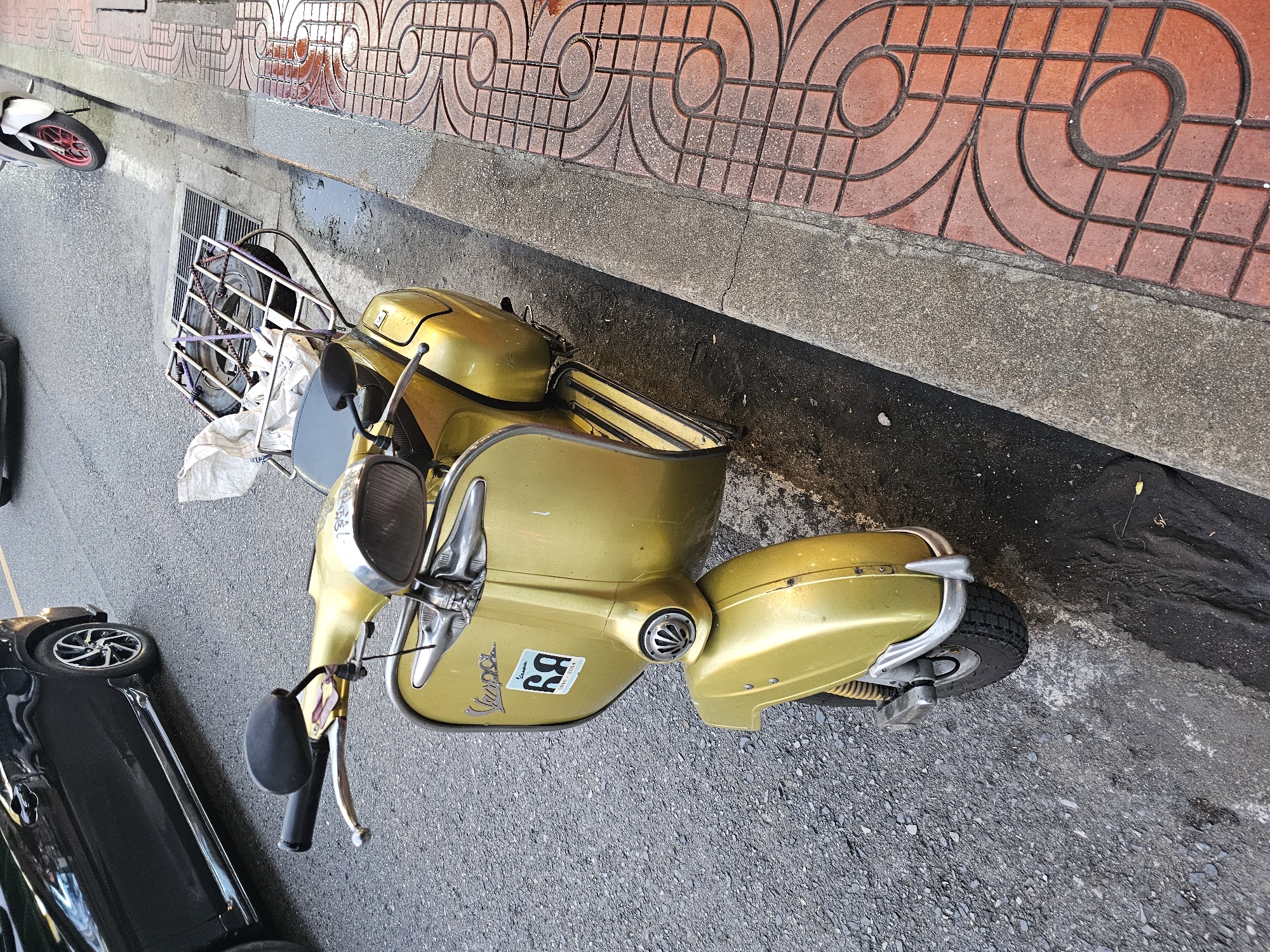 The image shows a close-up, eye-level view of a vintage gold Vespa scooter parked on a street in Bangkok, Thailand. The scooter is positioned facing slightly to the right, with its front wheel on the asphalt road. To the right of the scooter is a concrete curb and a sidewalk paved with red bricks featuring an intricate circular pattern. The road surface appears wet in places, suggesting recent rain.
Dominating the foreground is the scooter itself. It has a polished gold finish, and the "Vespa" logo is visible on the front. A sticker with the number "69" and the text "1946-2014" is also affixed to the front. The handlebars are black, and a chrome mirror is attached to the left side. A metal rack, possibly for carrying luggage, is mounted behind the seat, with a white bag and some other items secured to it. The front fender and headlight are also clearly visible.
In the background, a black car is partially visible on the left, parked on the same road. Further down the street, another scooter, white with a red wheel, is parked on the sidewalk. The environment appears to be an urban street scene, typical of Bangkok, with the presence of cars, scooters, and a well-maintained sidewalk. There are no people visible in the image. The lighting suggests it is daytime, possibly overcast or late afternoon given the shadows.